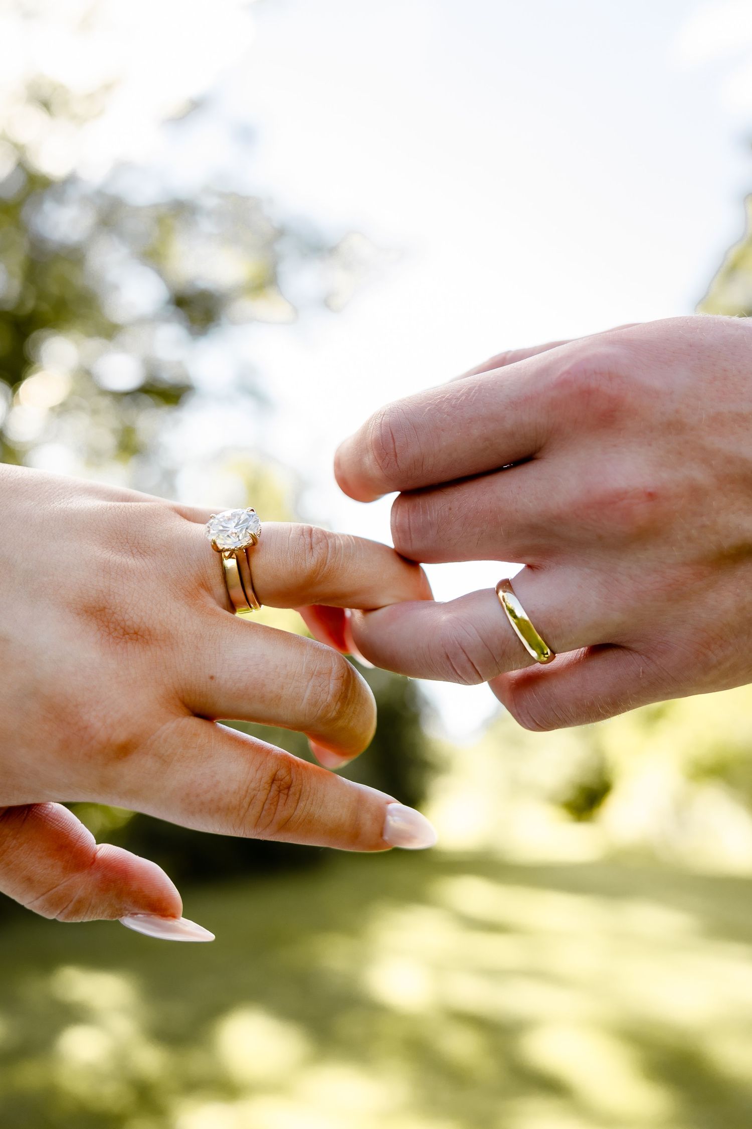 Two hands wearing gold wedding rings touch gently outdoors, symbolizing a romantic marriage commitment.