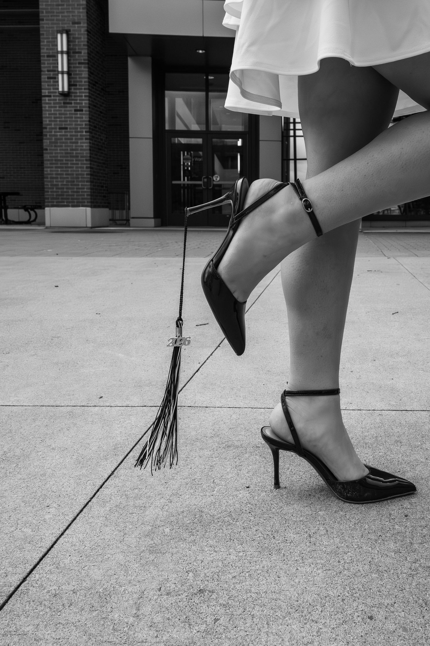 Black and white photo of woman's legs in strappy heels holding a tassel on sidewalk outside brick building.