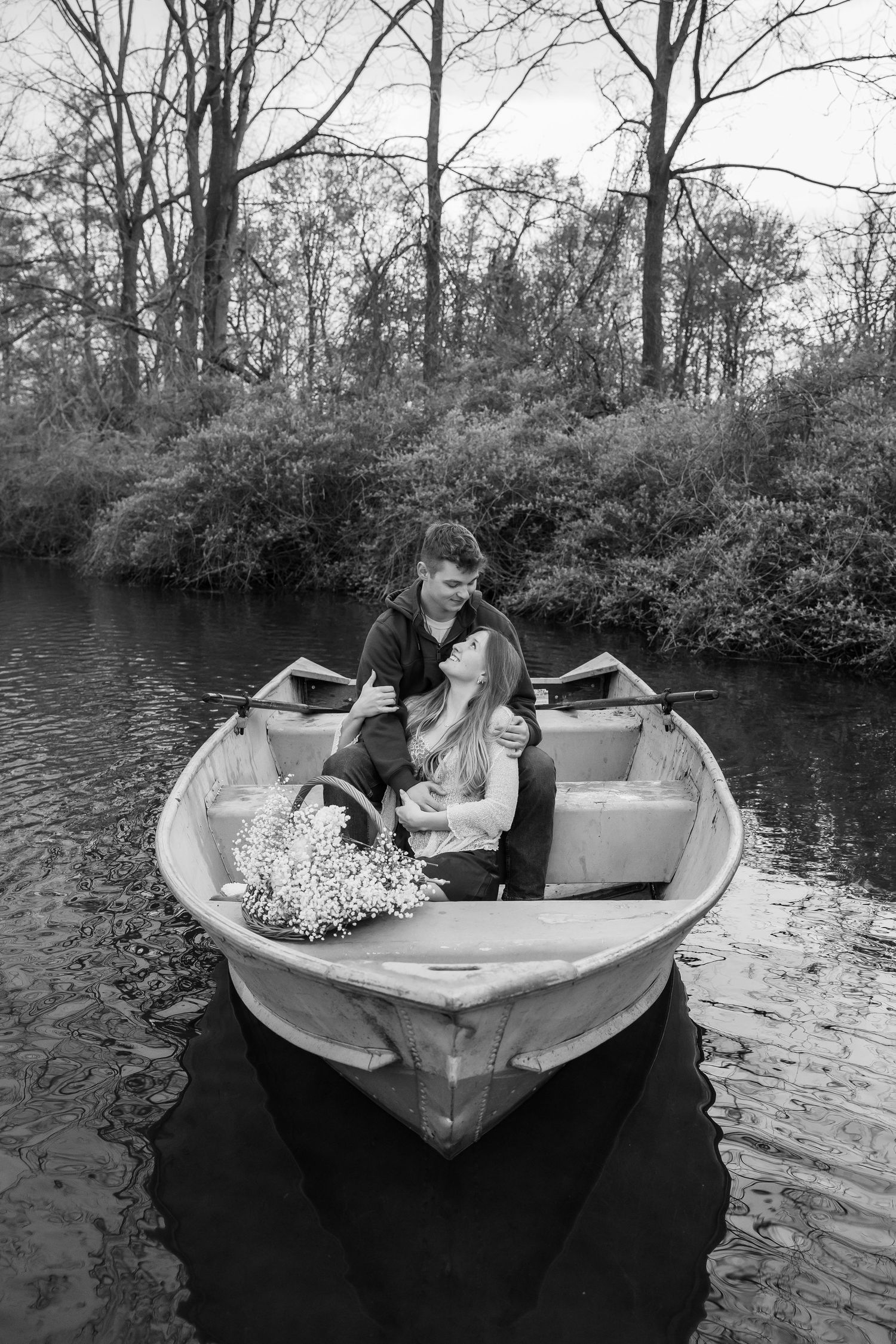 Black and white photo of a romantic couple embracing in a wooden rowboat on a calm river surrounded by trees.