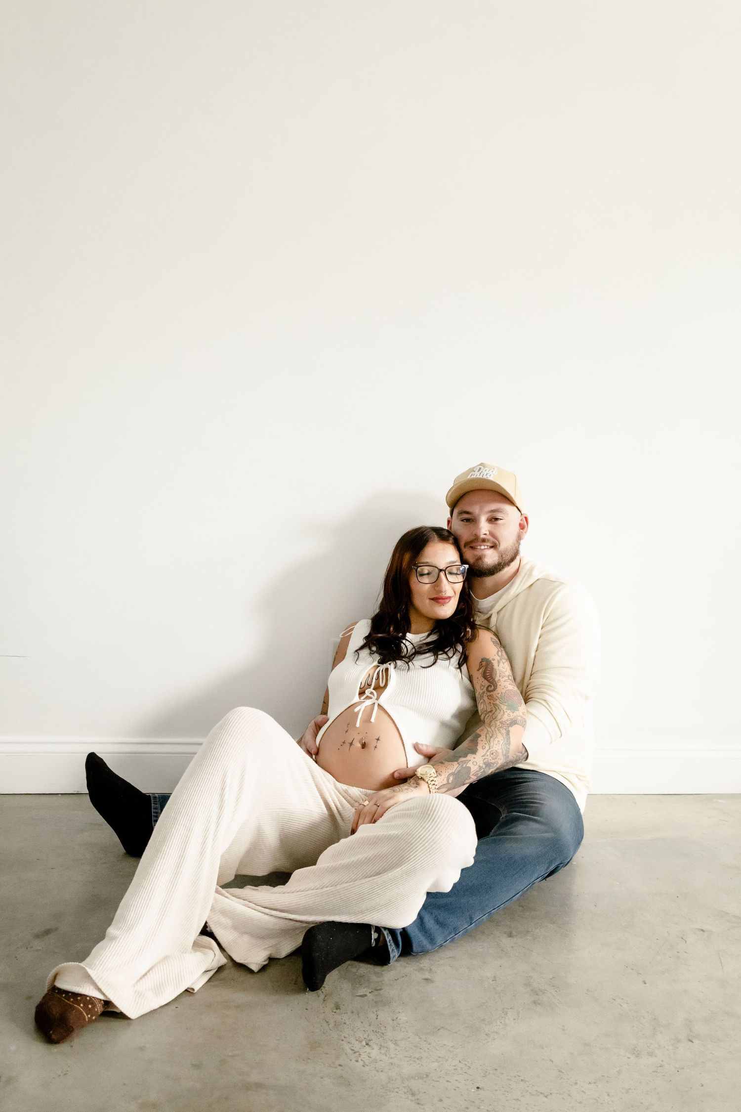 A pregnant couple sits on a concrete floor against a white wall, with the man embracing the woman from behind.