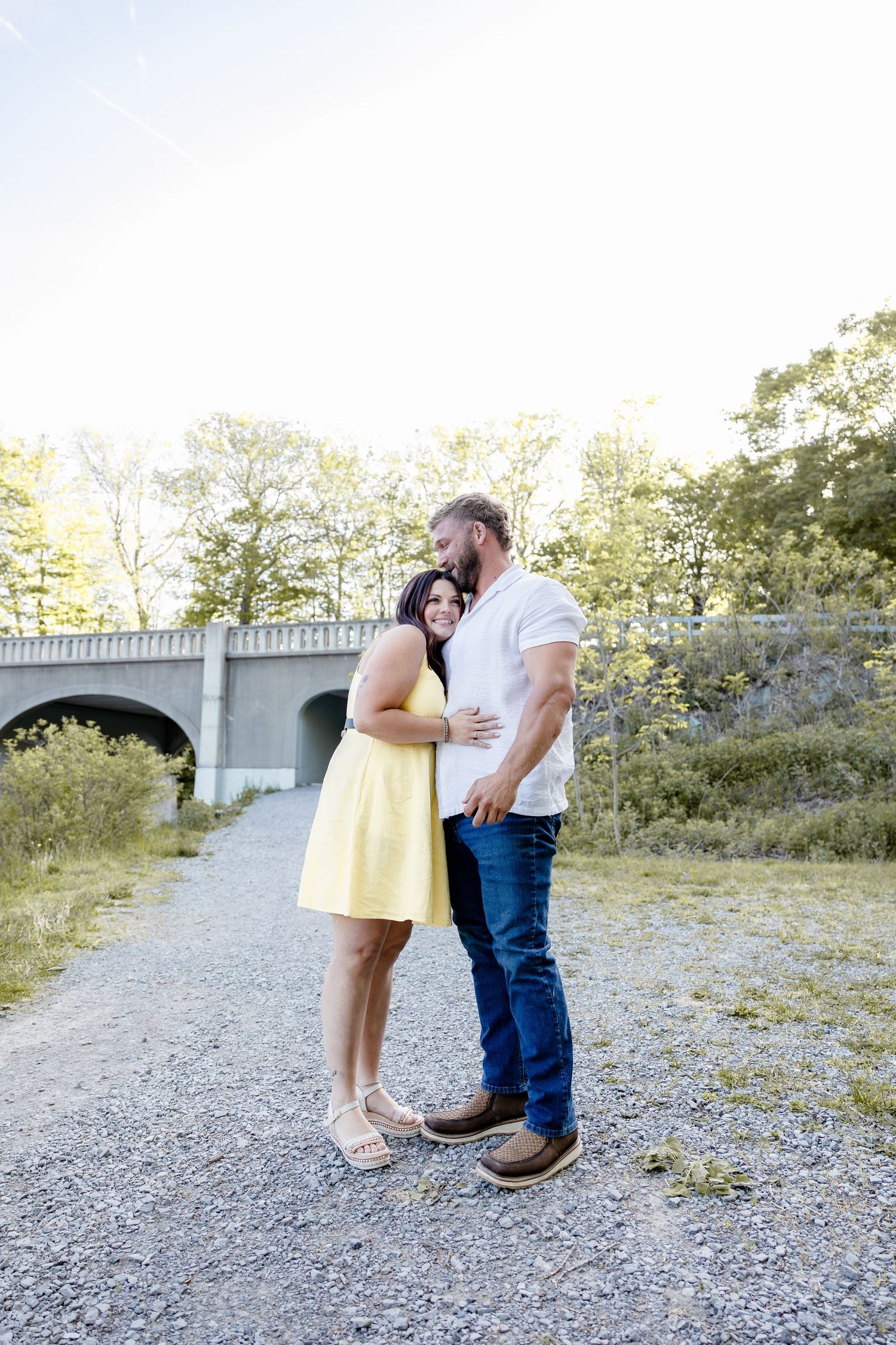 Couple embracing on gravel path near stone bridge, woman in yellow dress, man in white shirt and jeans, surrounded by trees.