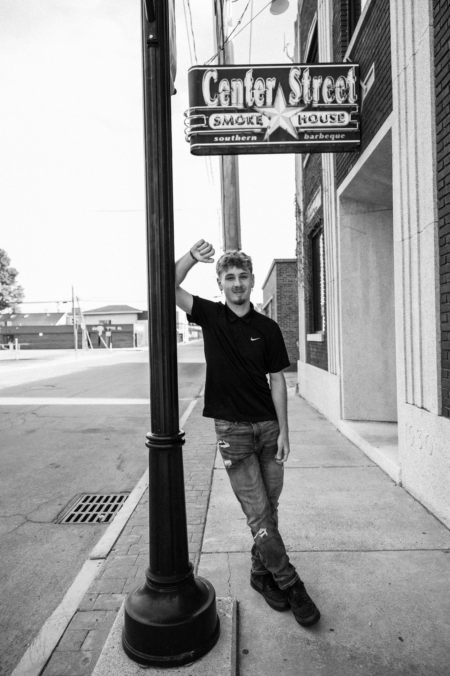 Young man in black Nike shirt and ripped jeans posing by Center Street Smokehouse sign on urban sidewalk.