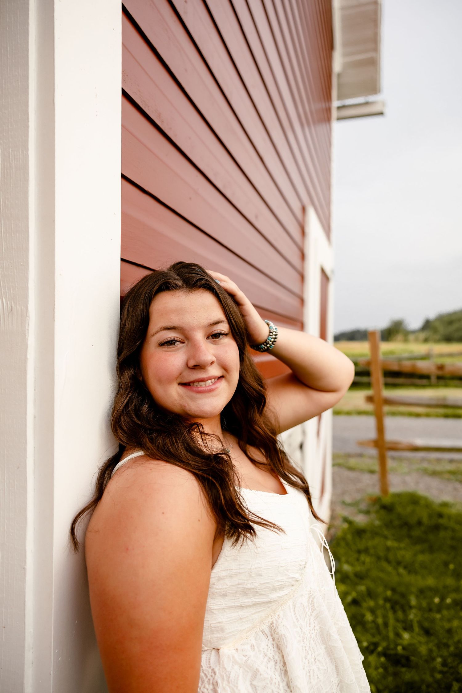 Young woman in white dress leaning against red barn wall, smiling outdoors on a farm.
