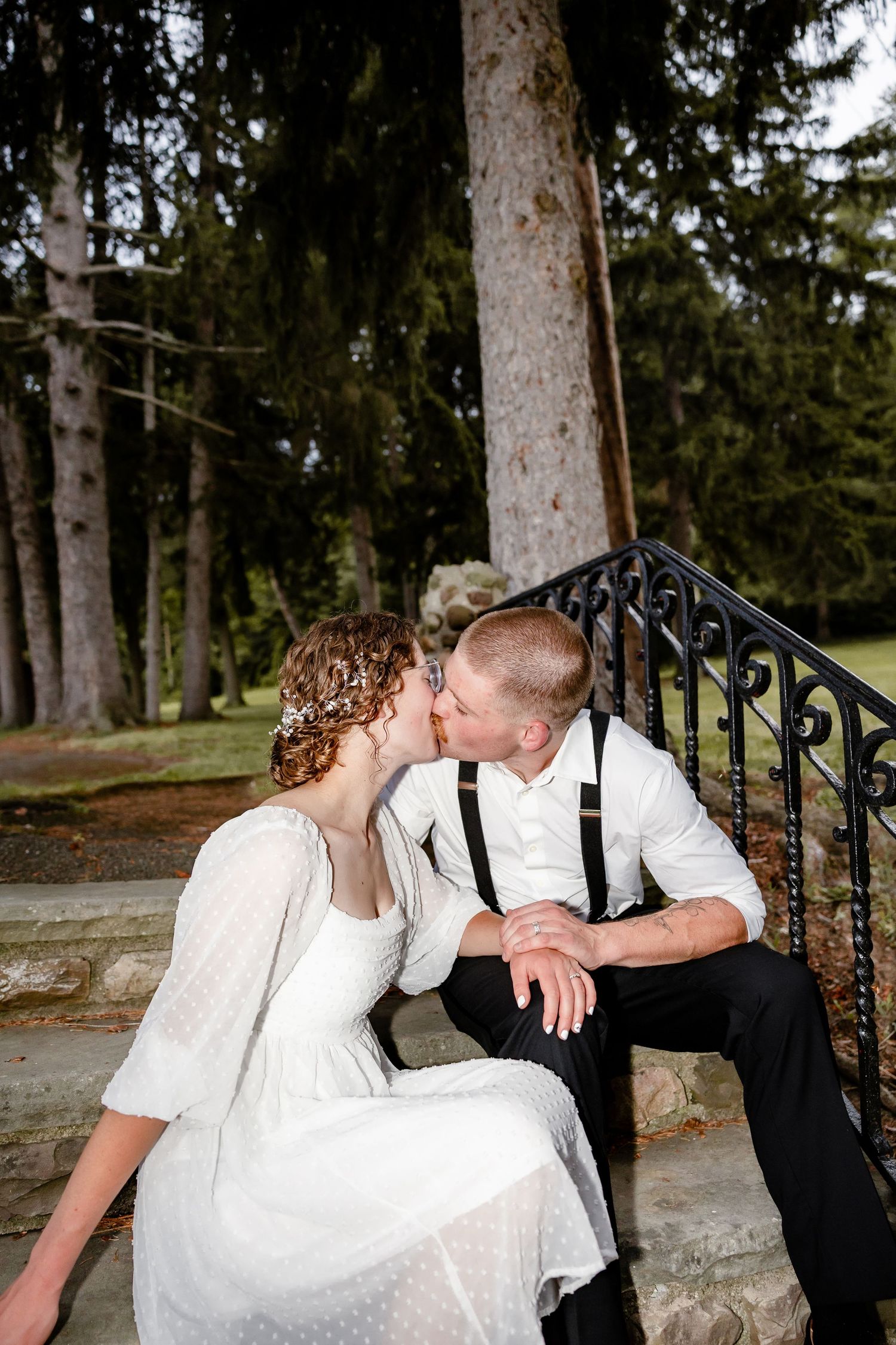 Bride and groom share a kiss while sitting on stone steps surrounded by tall evergreen trees.