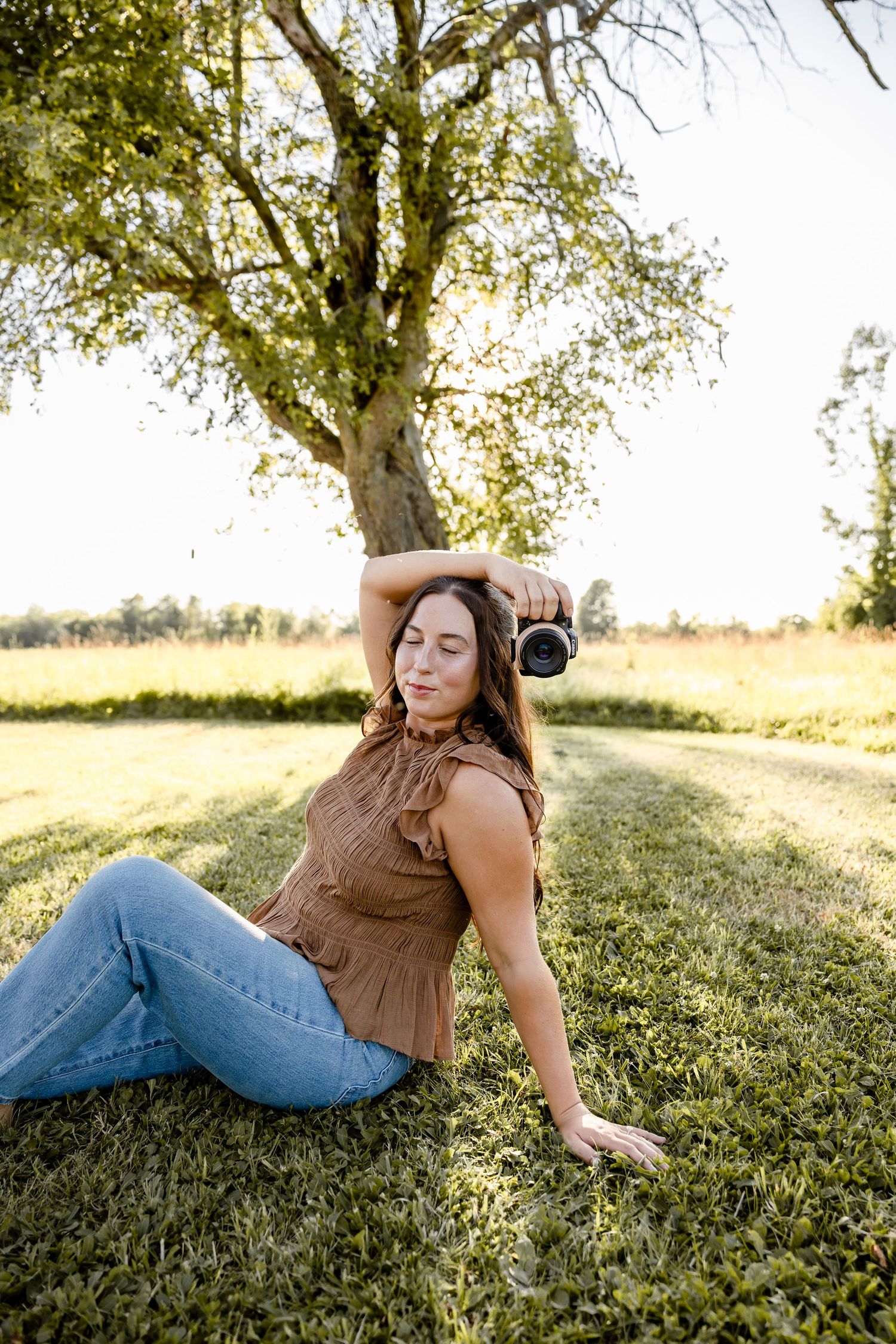 Woman in brown top and jeans sits on grass holding camera, relaxing under a tree in a sunny field.