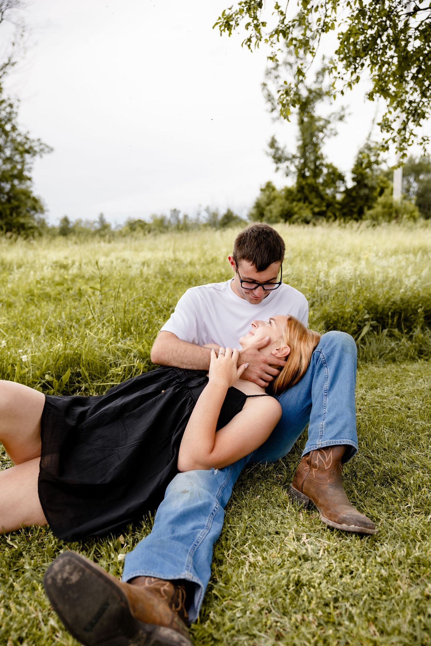 Couple relaxing in green field, woman lying in man's lap, both wearing jeans and boots, surrounded by lush trees.