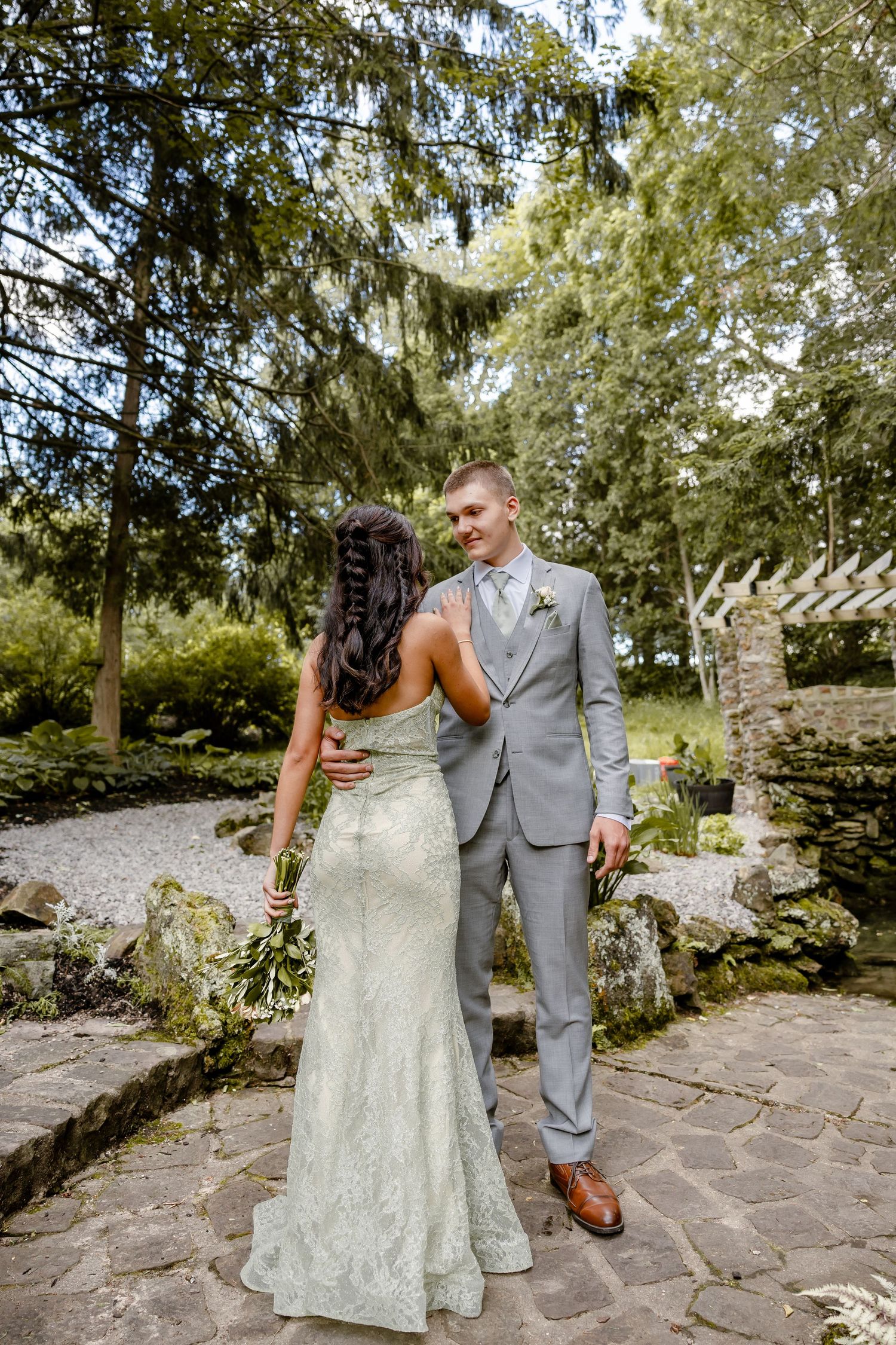 A bride in a mint lace gown and groom in gray suit share a romantic moment in a lush garden setting.