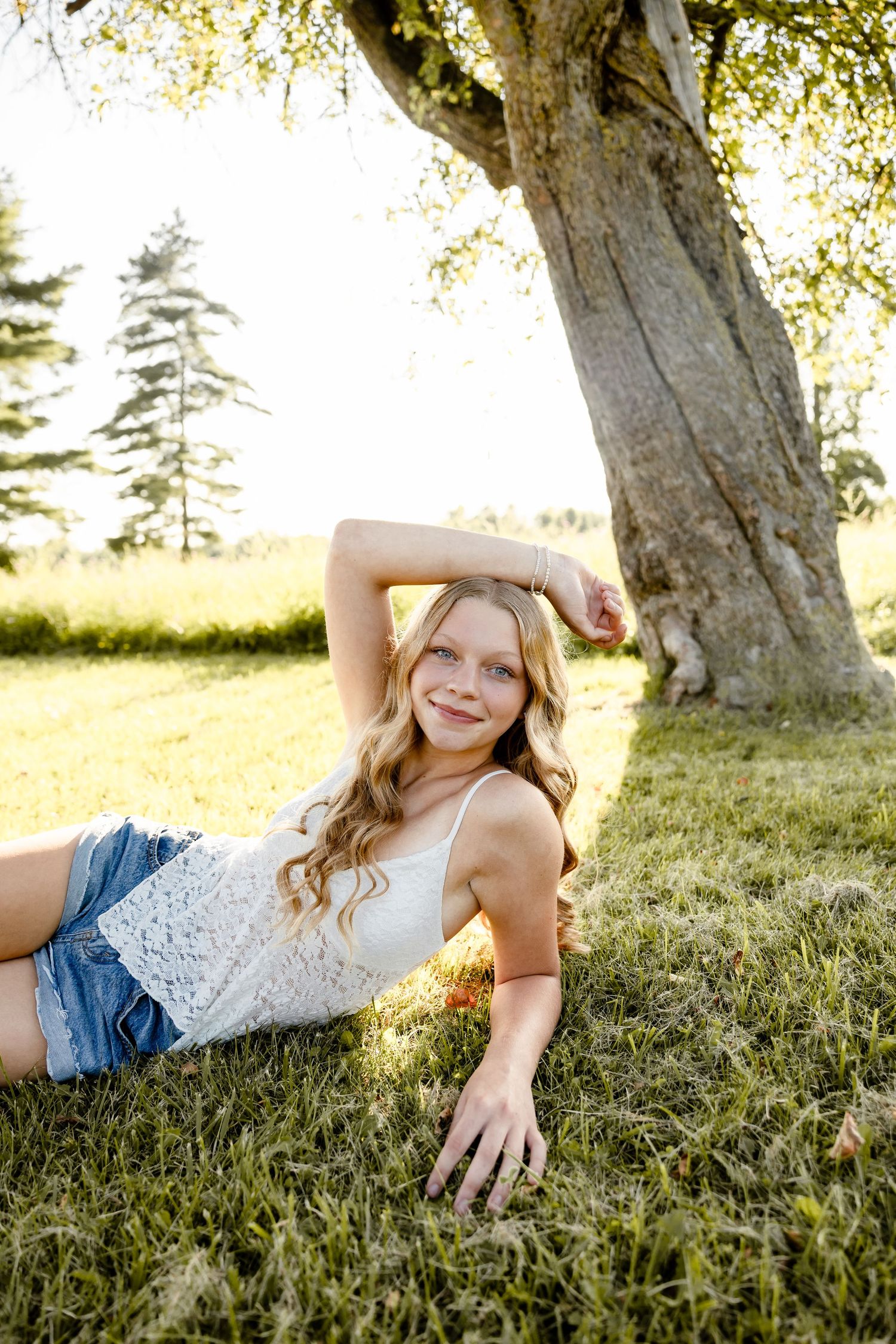 Young blonde girl in white lace top and denim shorts posing on grass beneath a large tree in golden sunlight.