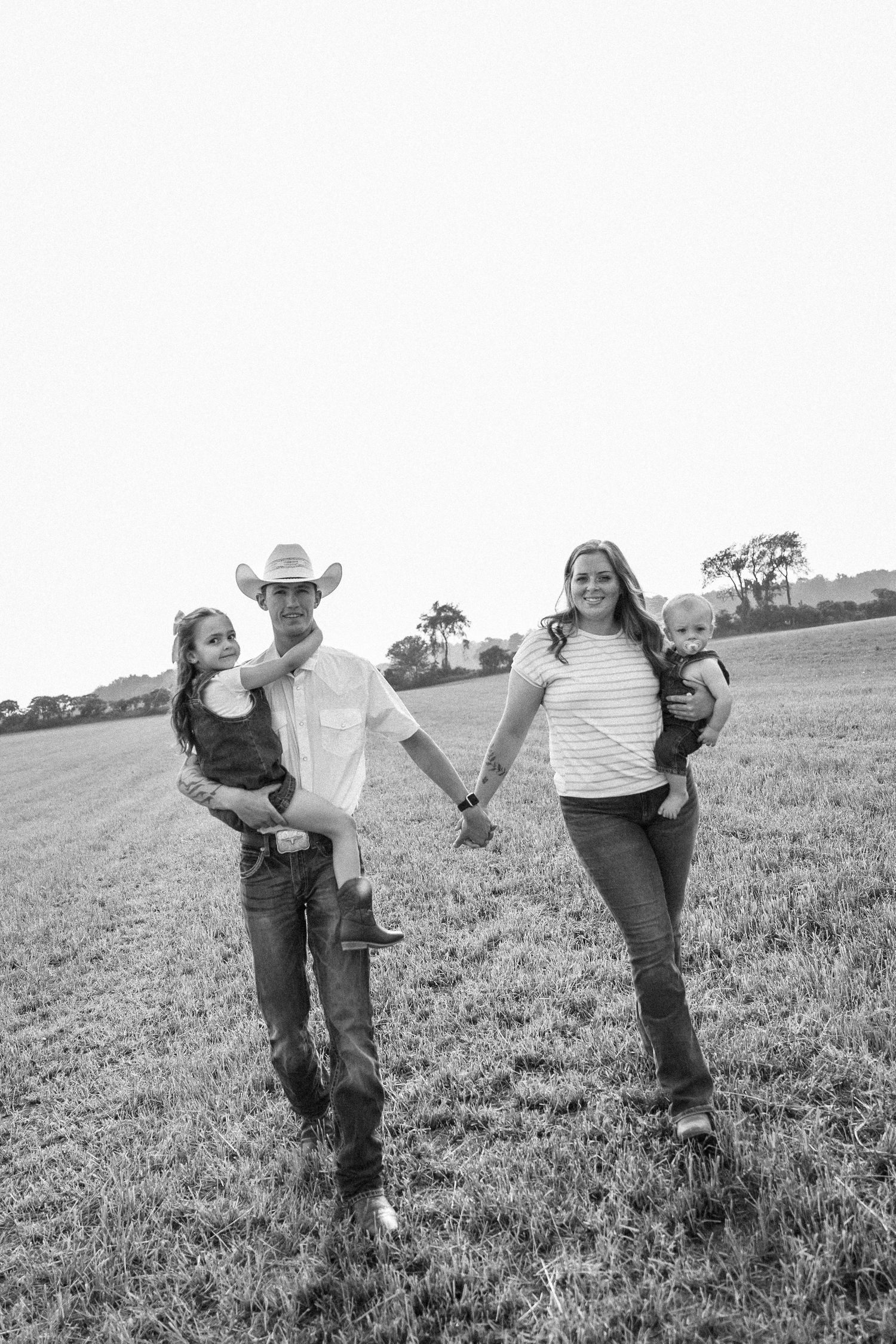 Black and white photo of a couple holding hands in a field, each carrying a child, man wearing cowboy hat and boots.