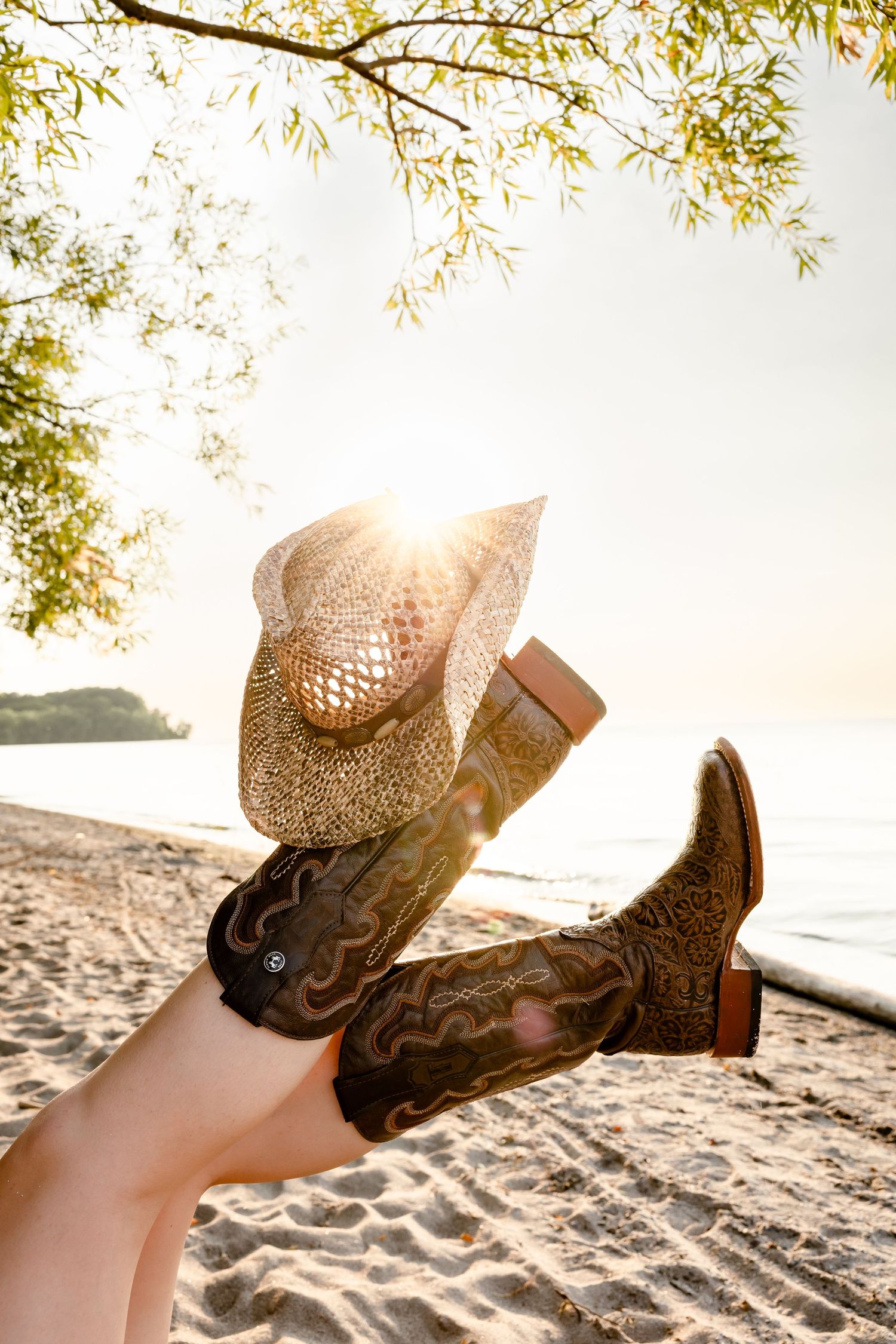 Person relaxing on sandy beach wearing cowboy boots and straw hat raised in the air with golden sunset light.