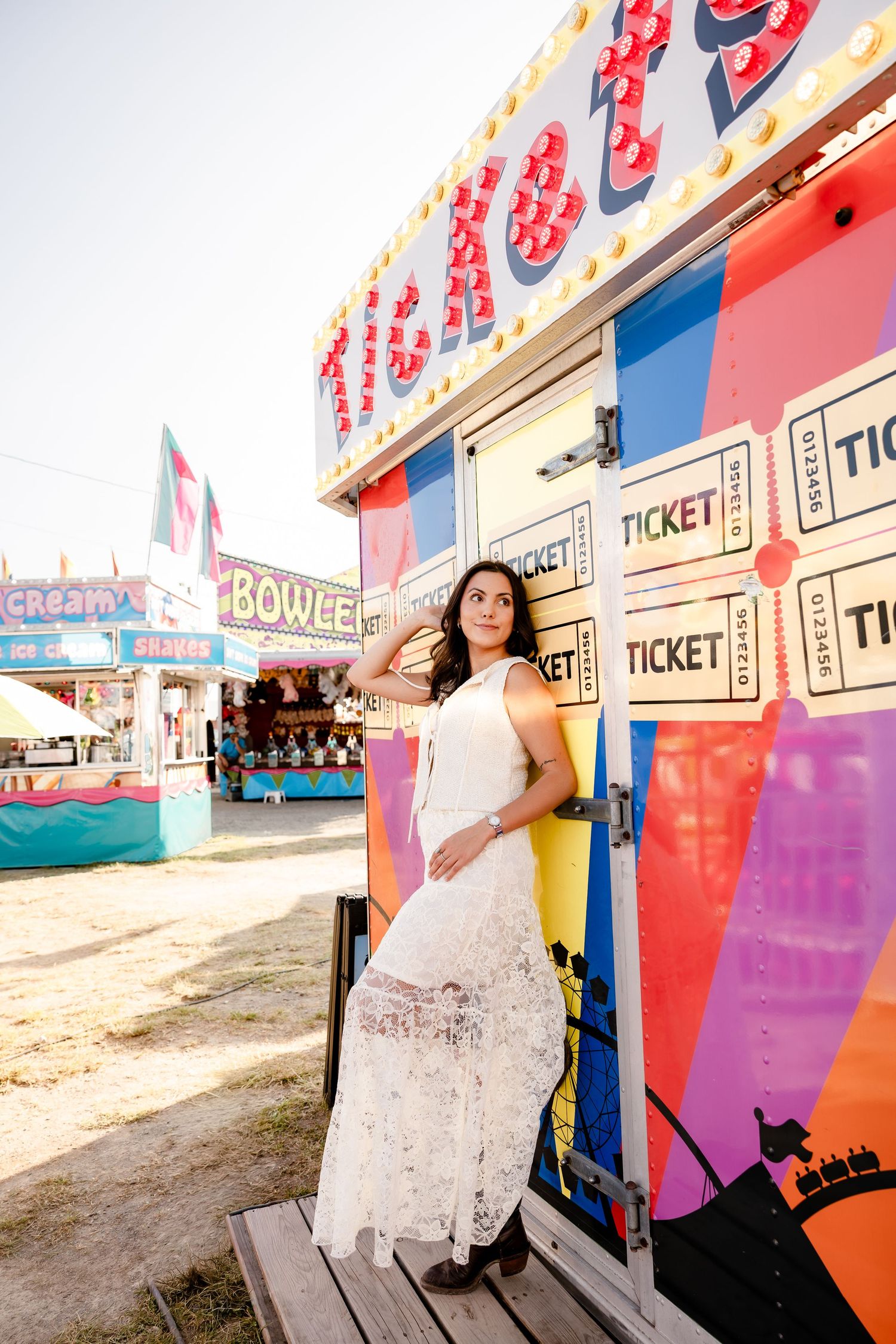 Young woman in a white lace dress posing by a colorful carnival ticket booth on a sunny day.
