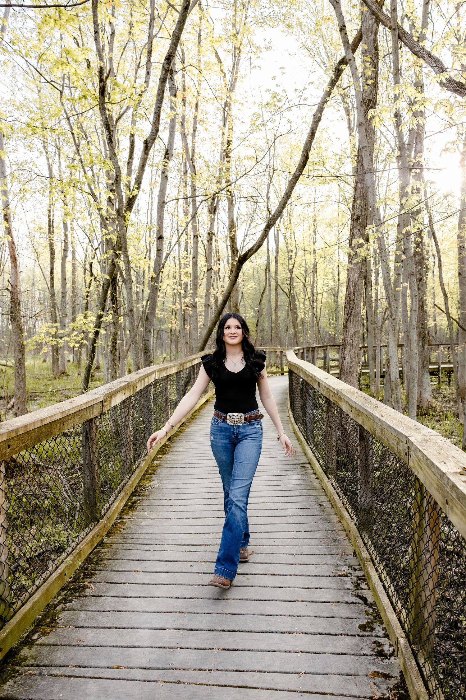 Young woman in black top and jeans walks confidently on a wooden forest boardwalk surrounded by spring trees.
