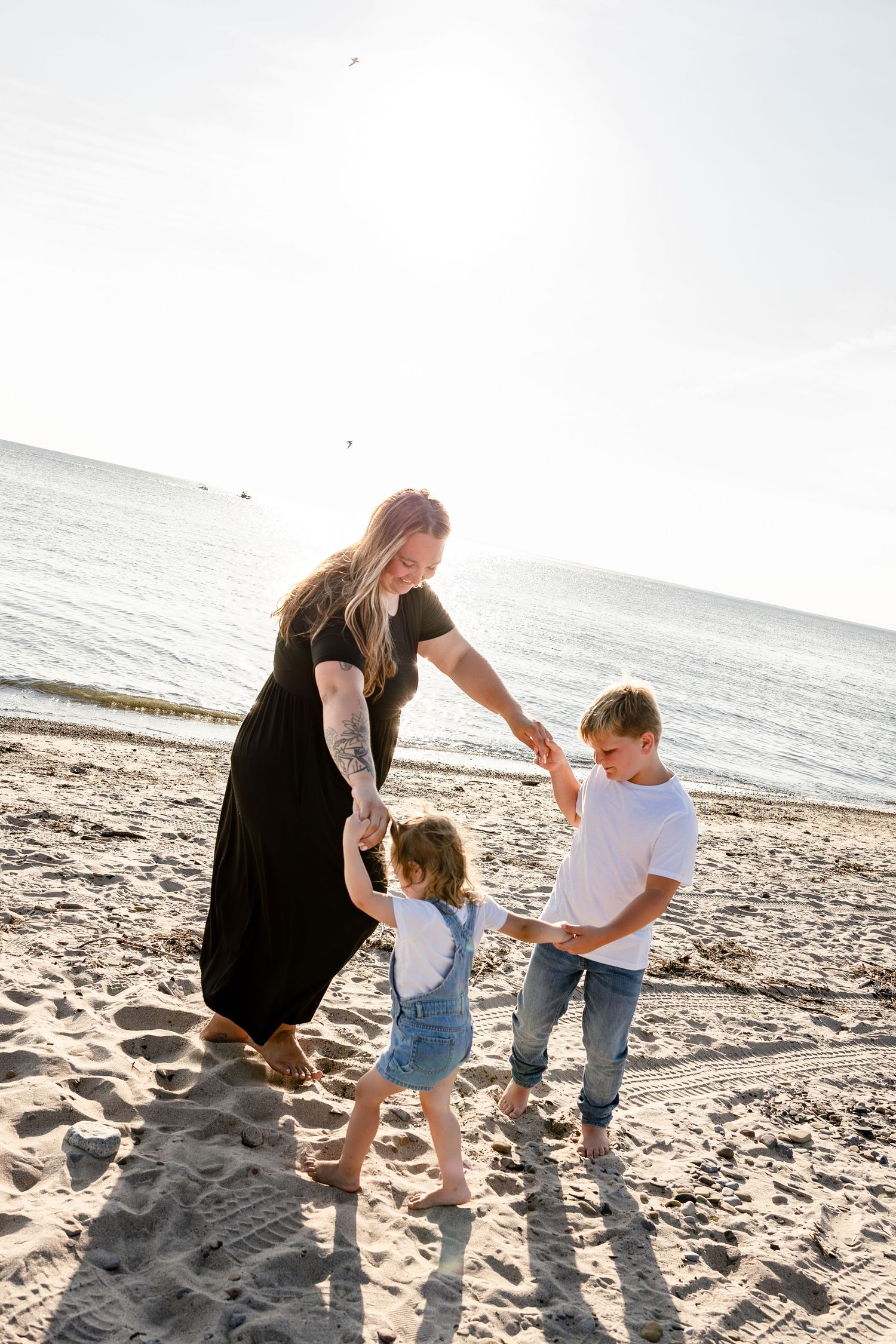 A woman in a black dress plays with two children on a sunny beach, holding hands near the ocean shoreline.