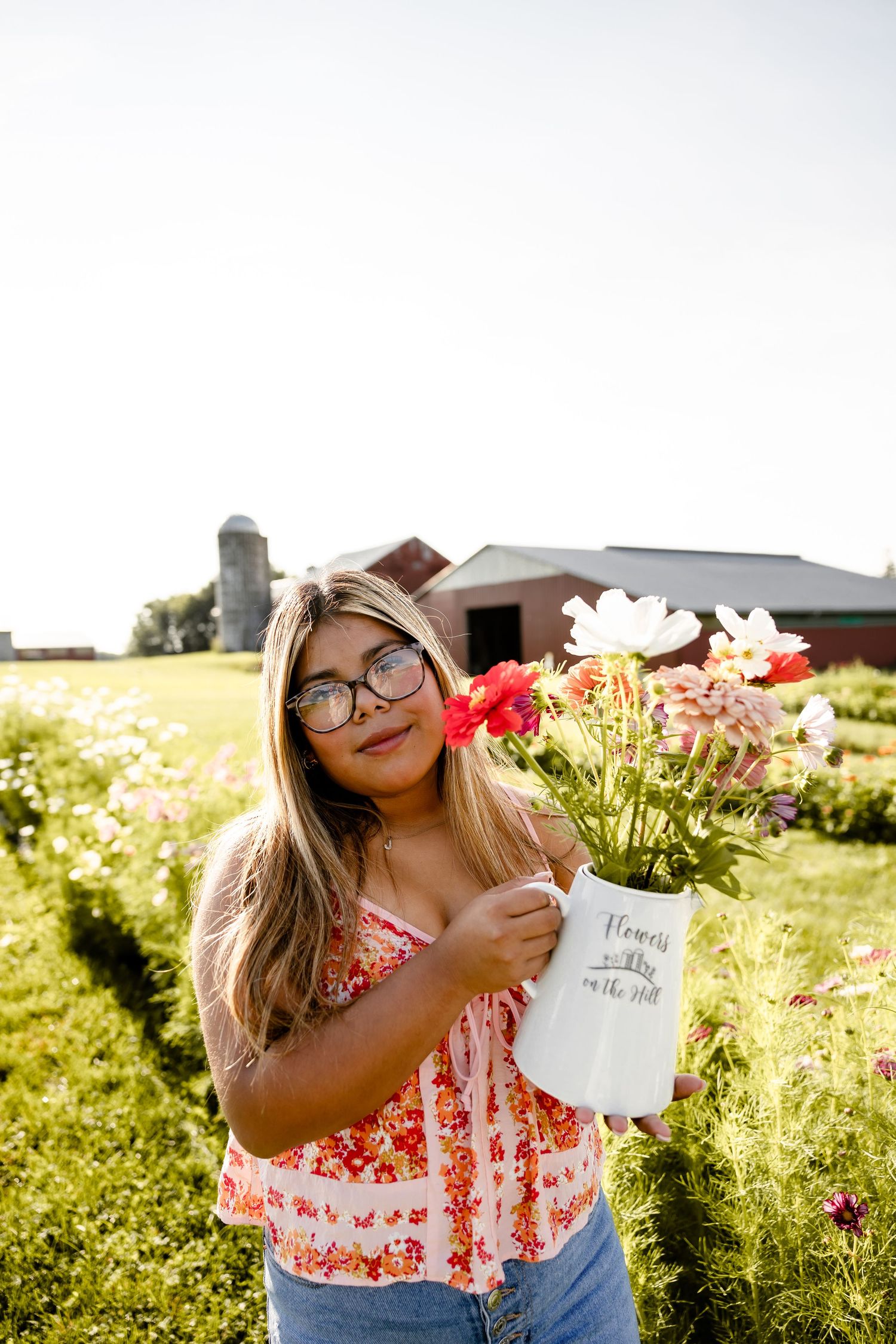 Young woman with glasses holding white pitcher of fresh picked flowers at a sunny flower farm.