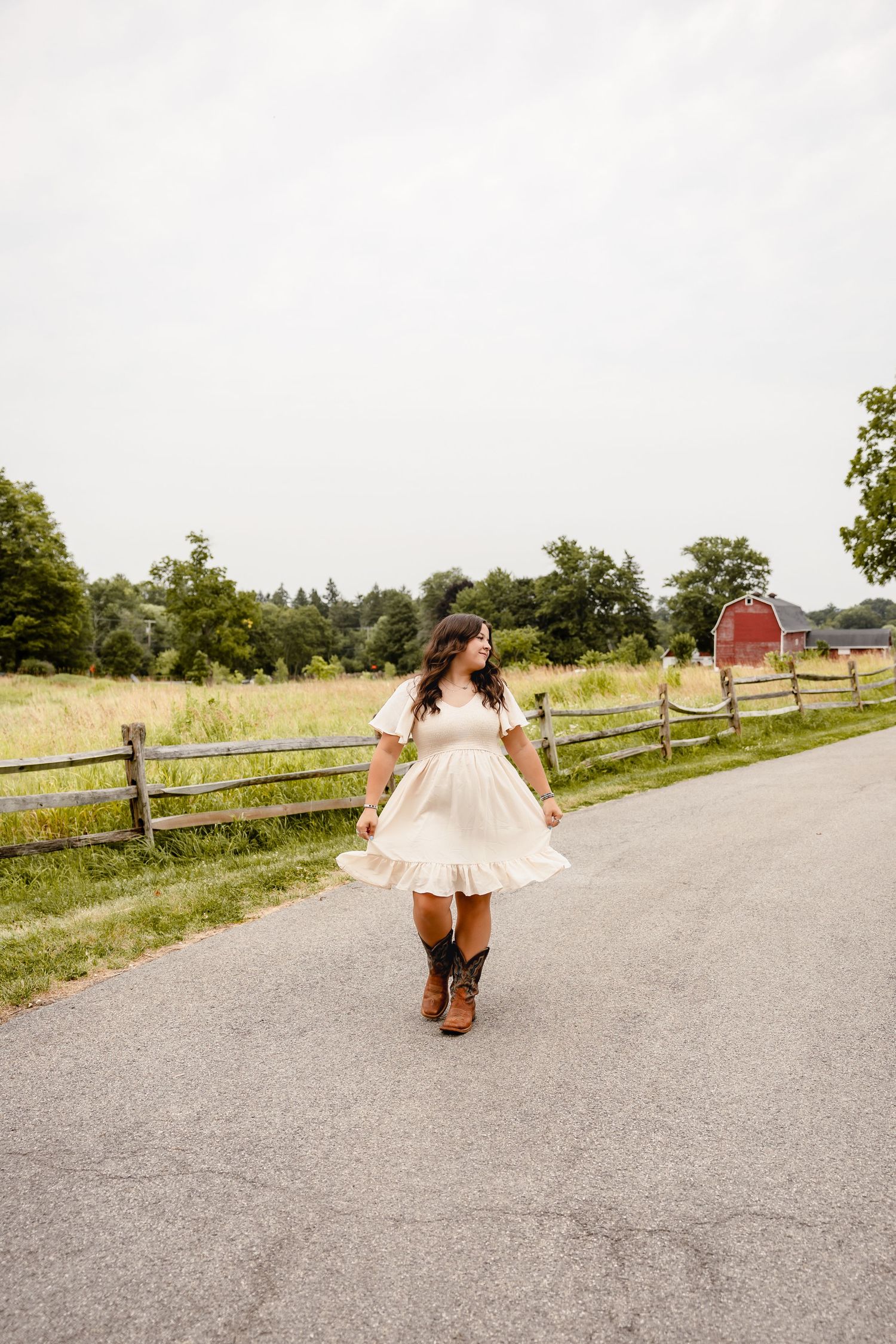 Woman in white dress and cowboy boots walking on a rural road with a red barn and wooden fence in background.