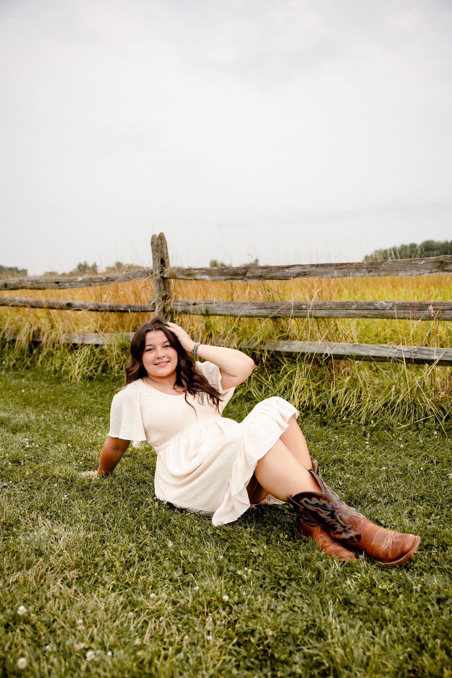 Woman in white dress and cowboy boots sitting on grass by rustic wooden fence in a golden field.