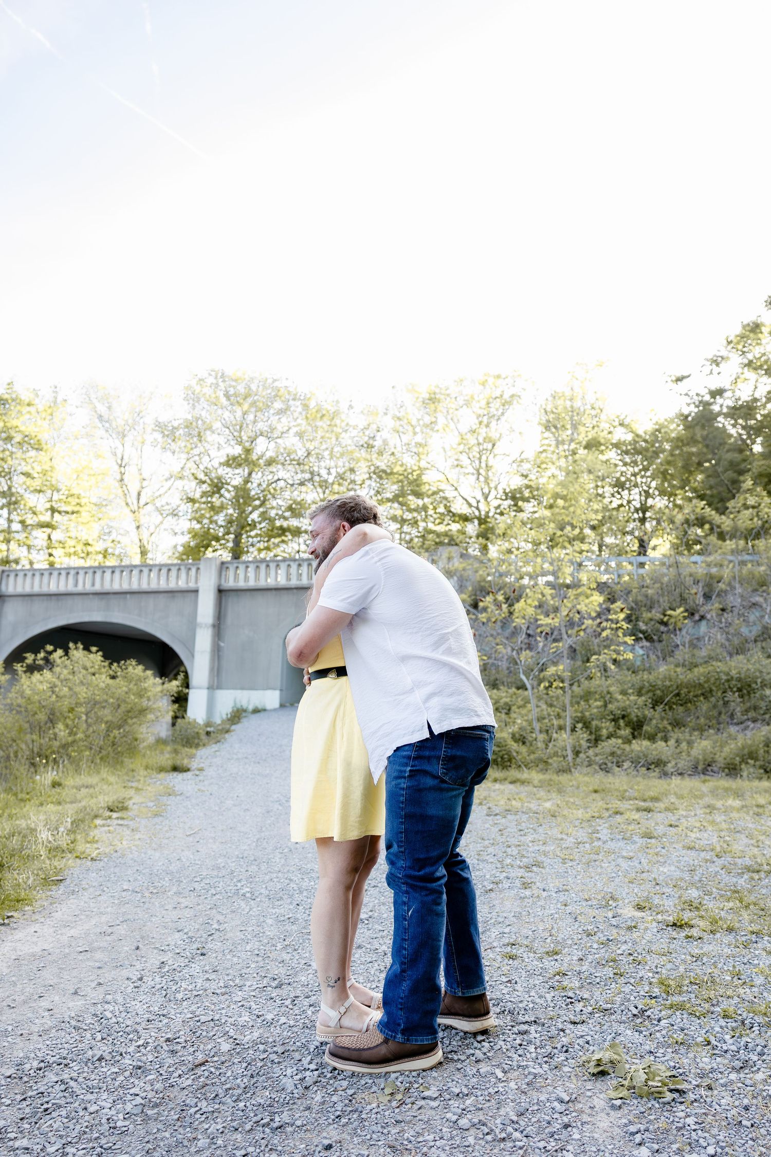 Couple embracing on a gravel path near a stone bridge surrounded by trees on a sunny day.