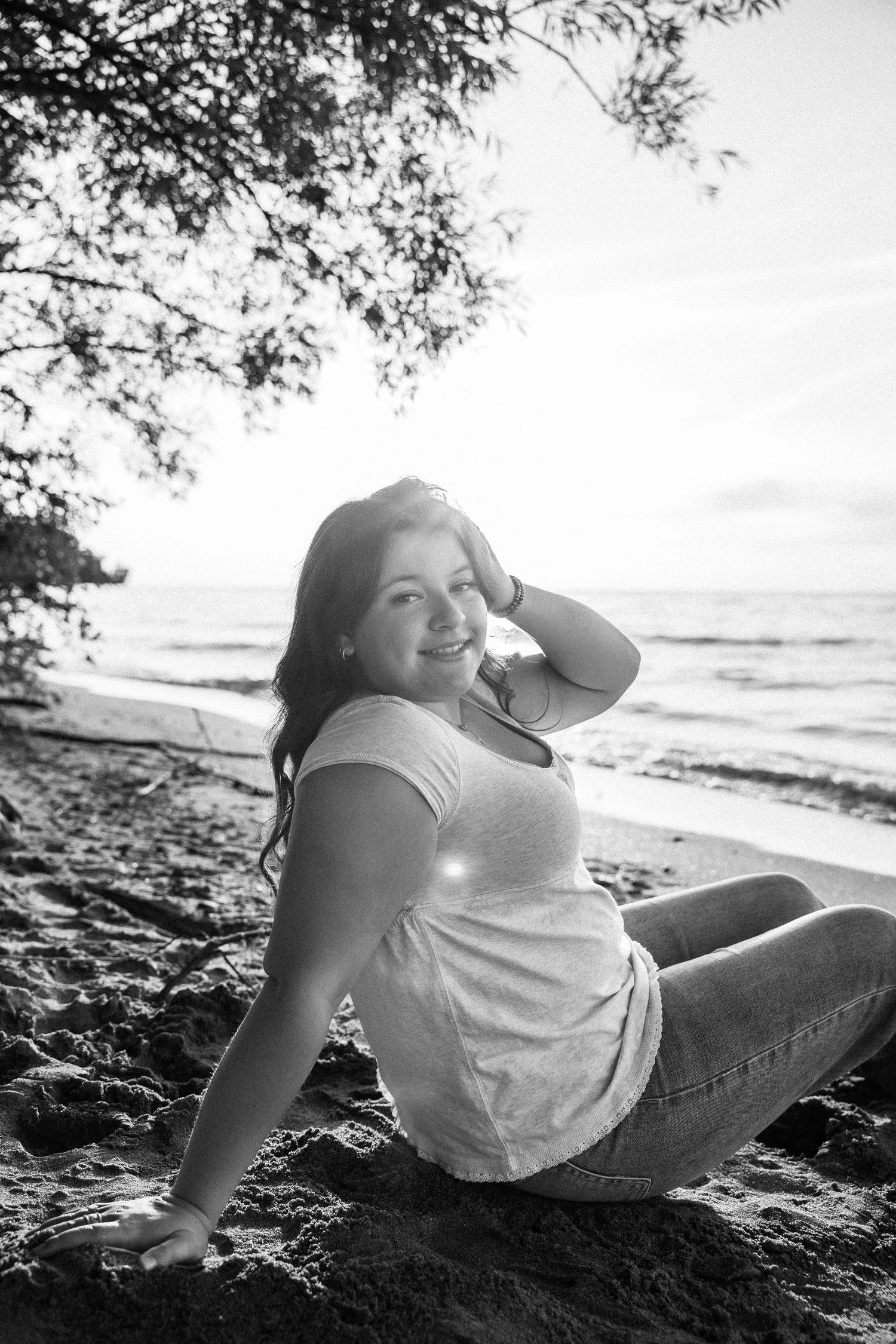 Black and white photo of a young woman sitting on a sandy beach, smiling with hand in hair, ocean in background.