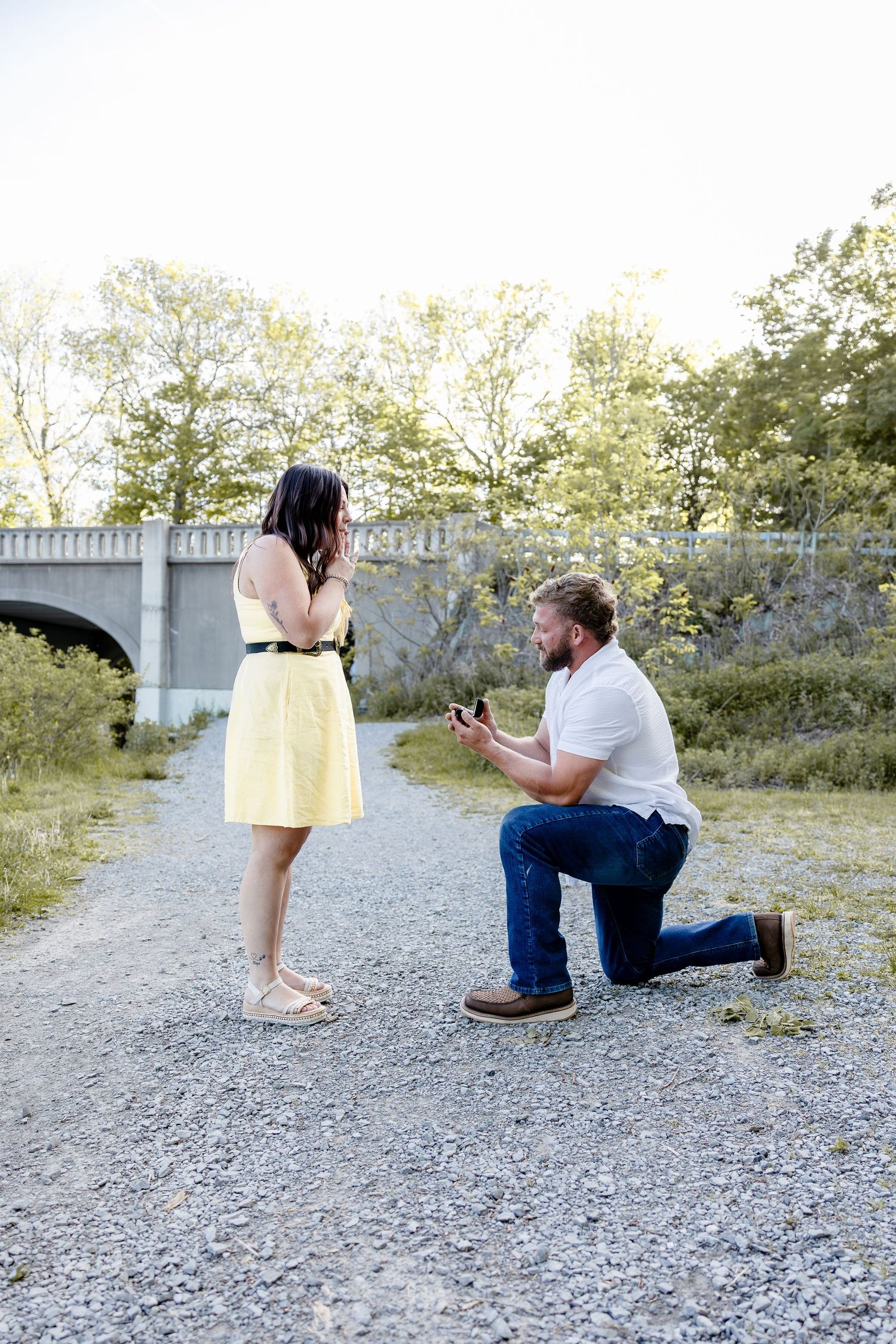 Man kneeling on one knee proposing to woman in yellow dress on gravel path surrounded by trees.