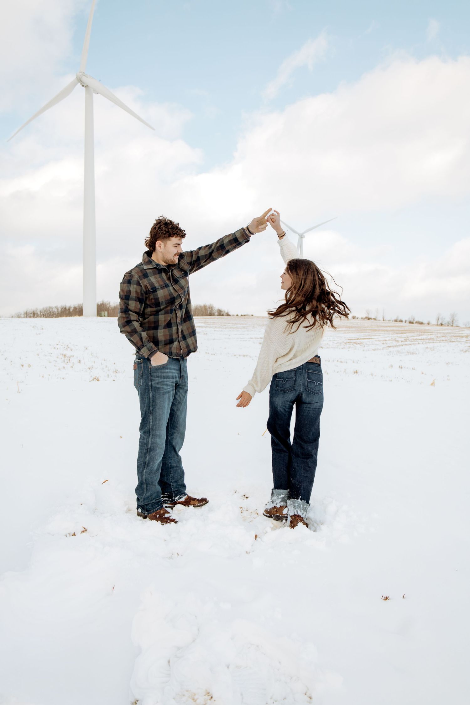 Couple dancing together in a snowy field with a wind turbine in the background on a cloudy winter day.