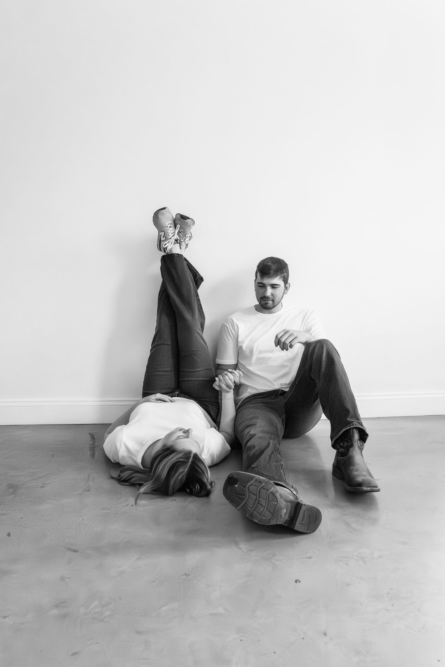 Black and white photo of a couple sitting on floor against white wall, woman lying down holding mans hand.