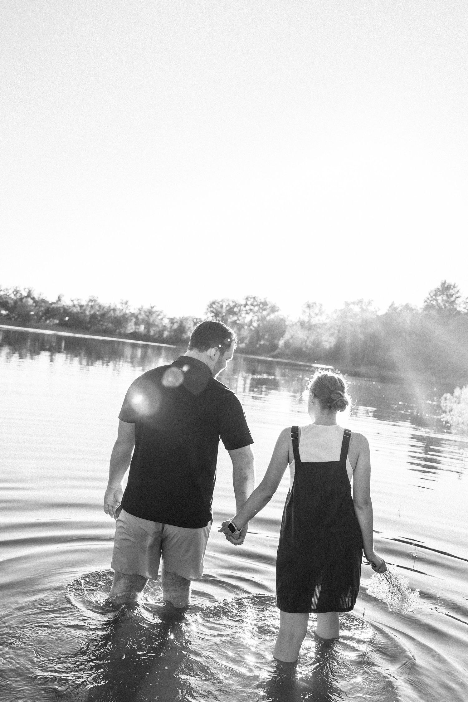 Black and white photo of a couple holding hands wading in a lake at sunset.