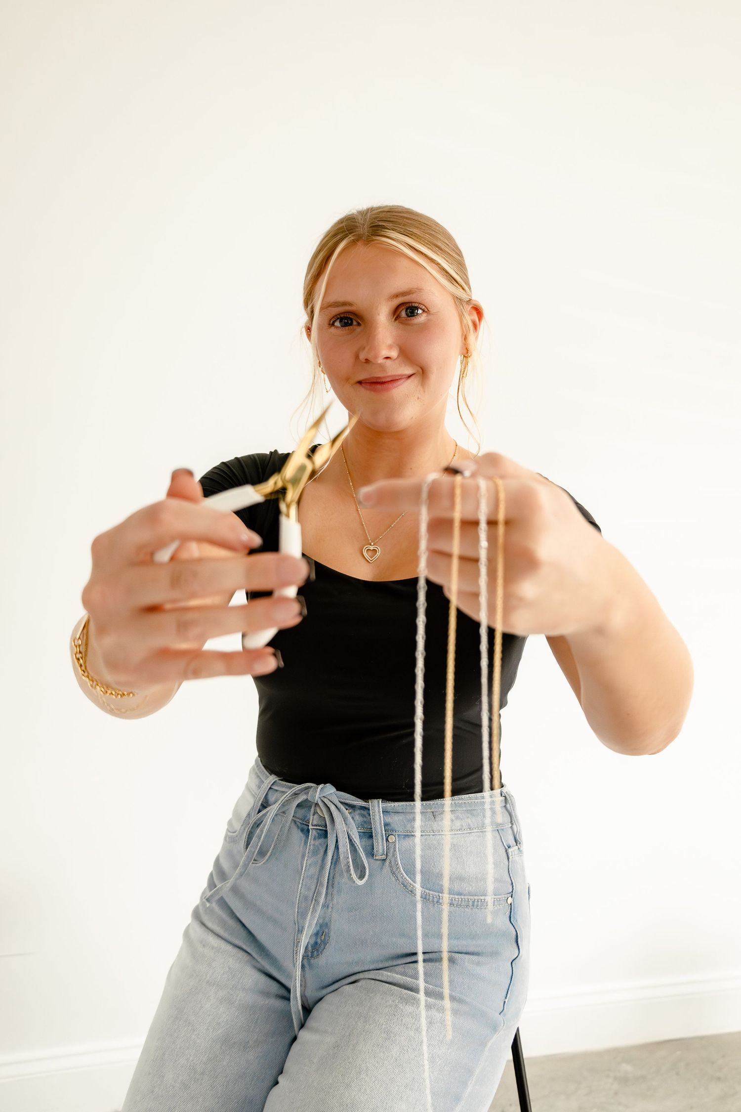 Smiling blonde woman in black top and jeans cutting gold chain necklaces with gold scissors.