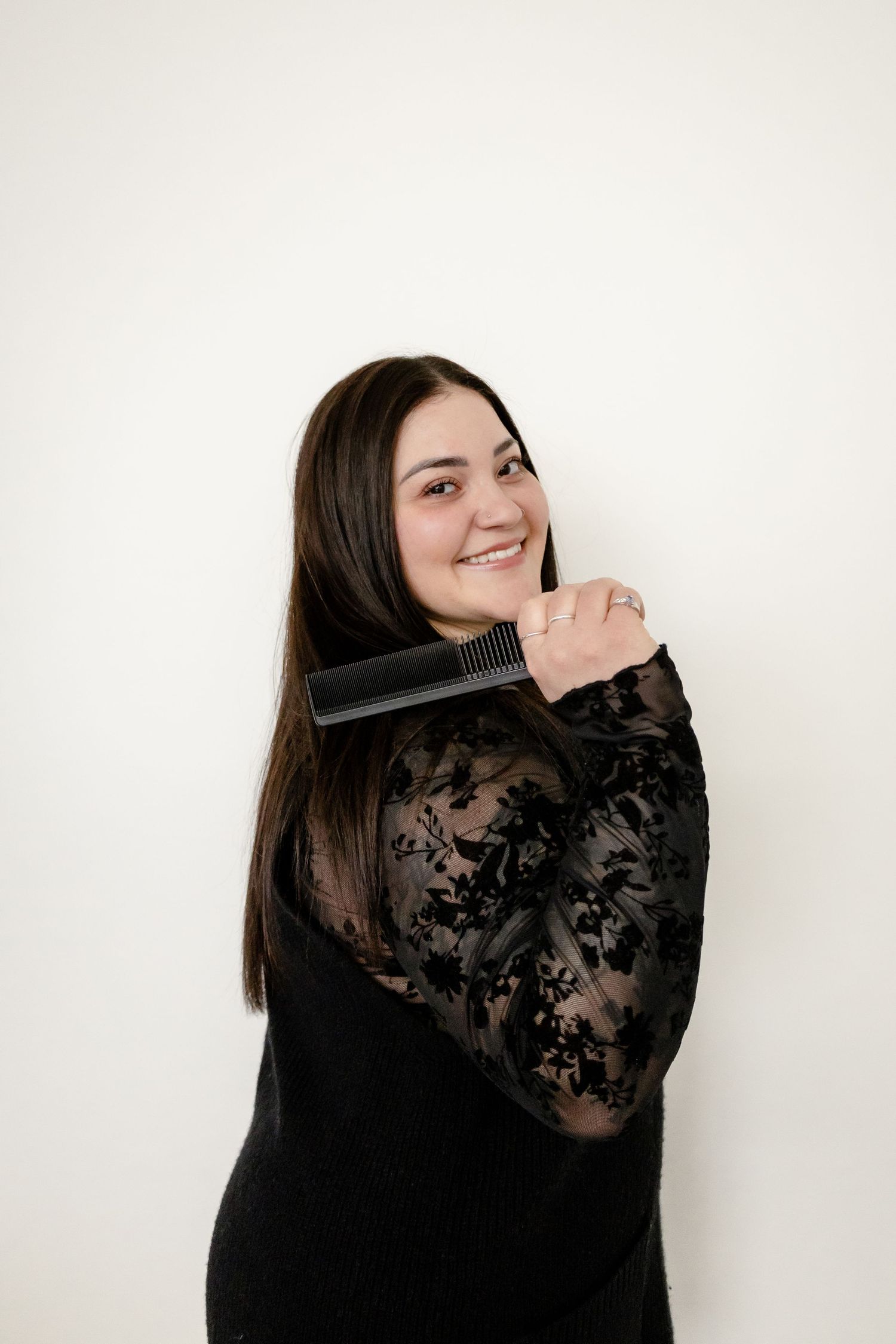 Smiling brunette woman in black lace top brushing her long straight hair against a white background.