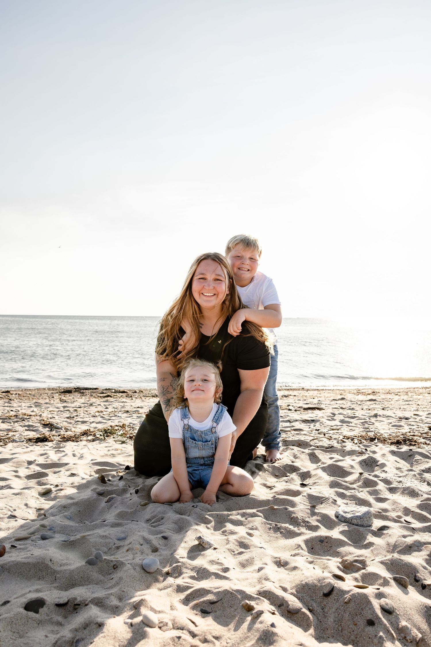 A smiling family of three poses on a sandy beach, with a mother and two children enjoying a sunny day by the ocean.