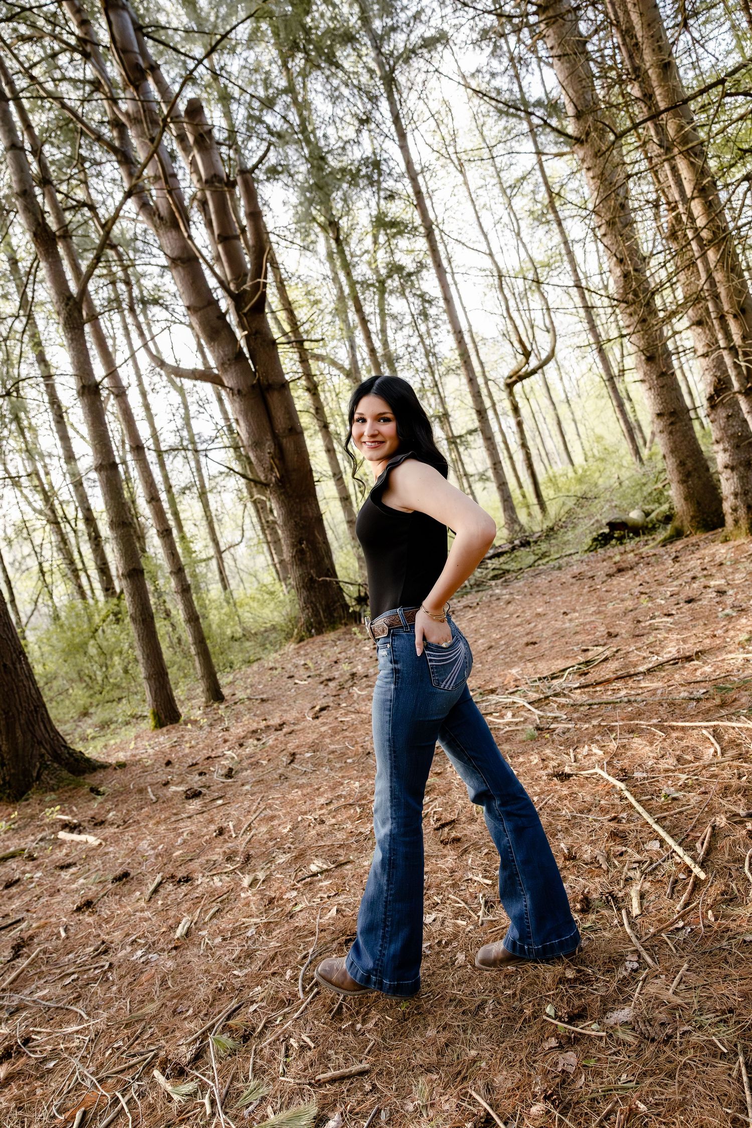 Young woman in black top and flare jeans posing confidently in a sunlit pine forest.