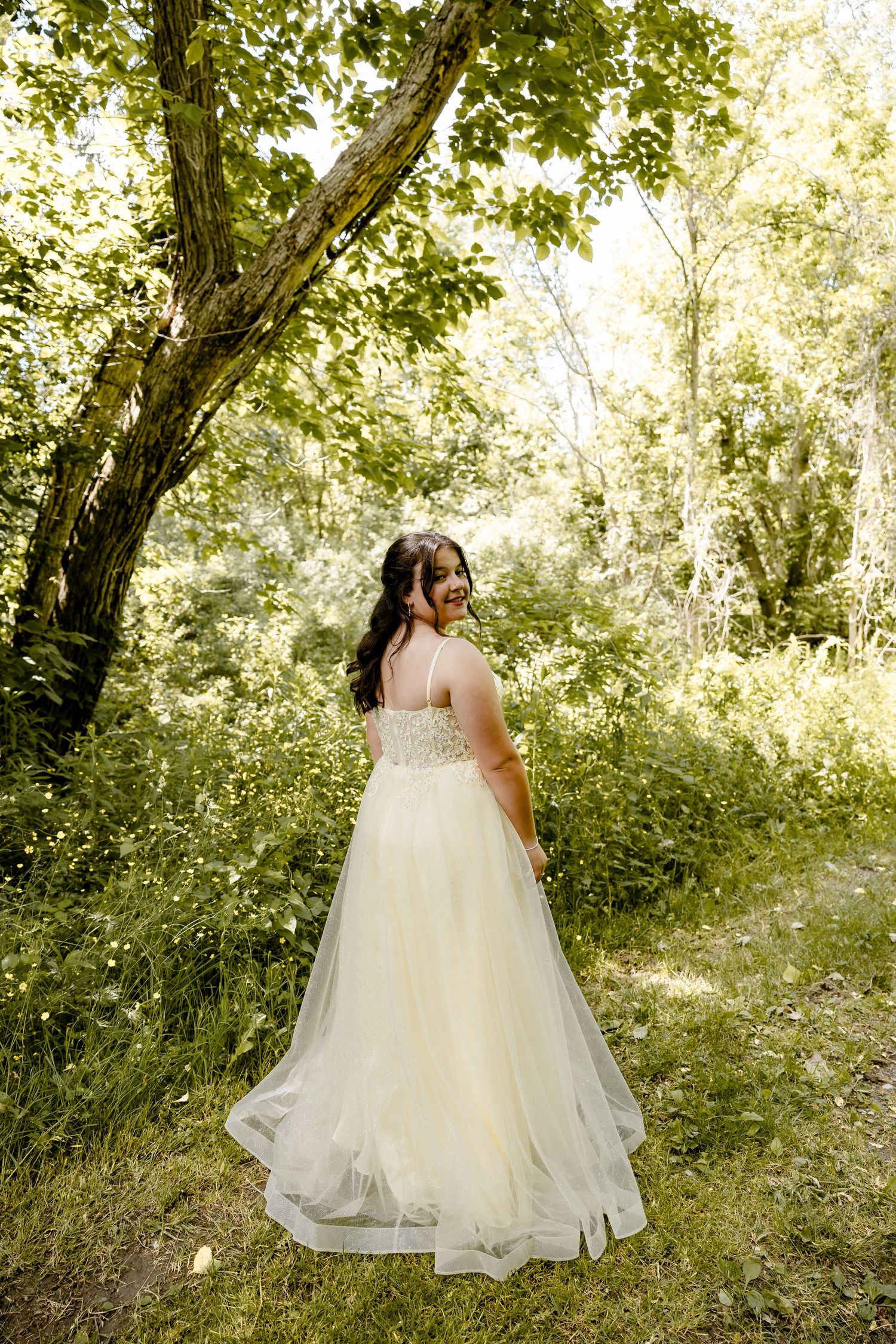 Bride in ivory tulle wedding gown standing in lush green forest, looking back over shoulder with a smile.