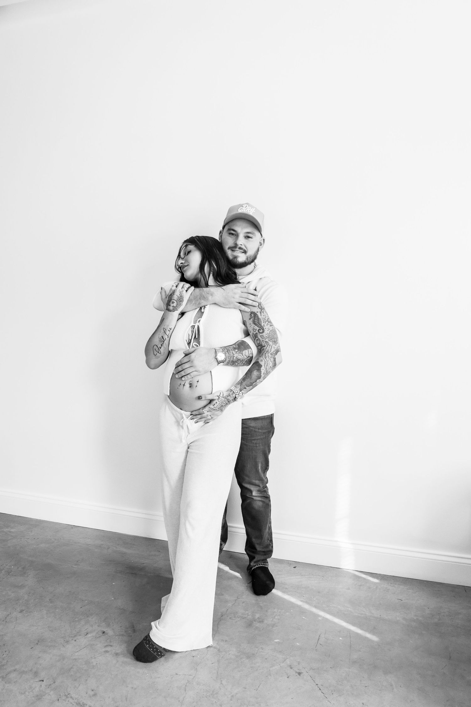 Black and white photo of a tattooed couple embracing, woman in white outfit with baby bump, man in cap hugging her from behind.