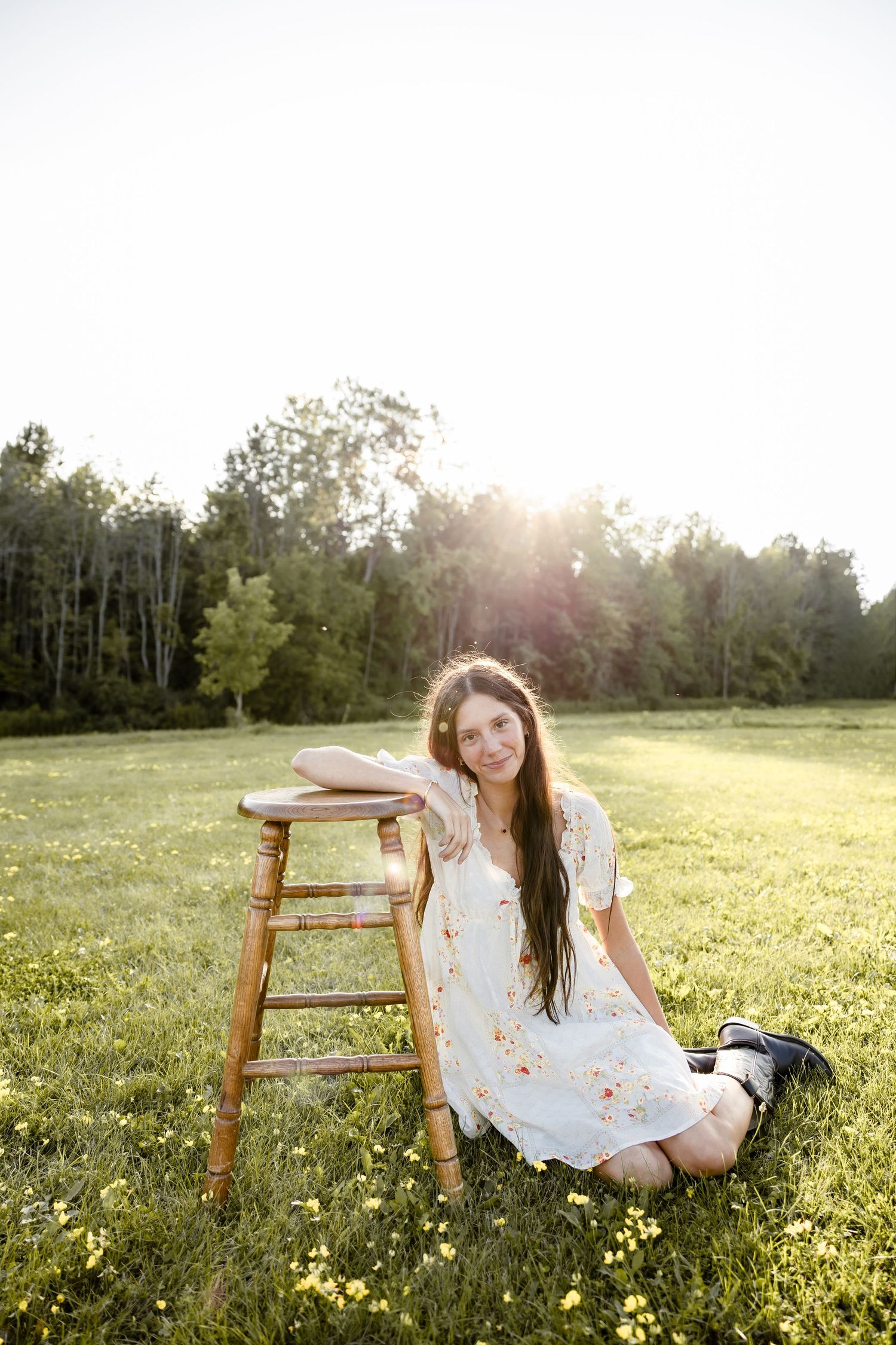 Young woman in floral dress sitting on grass field leaning on wooden stool with sunlit trees in background.