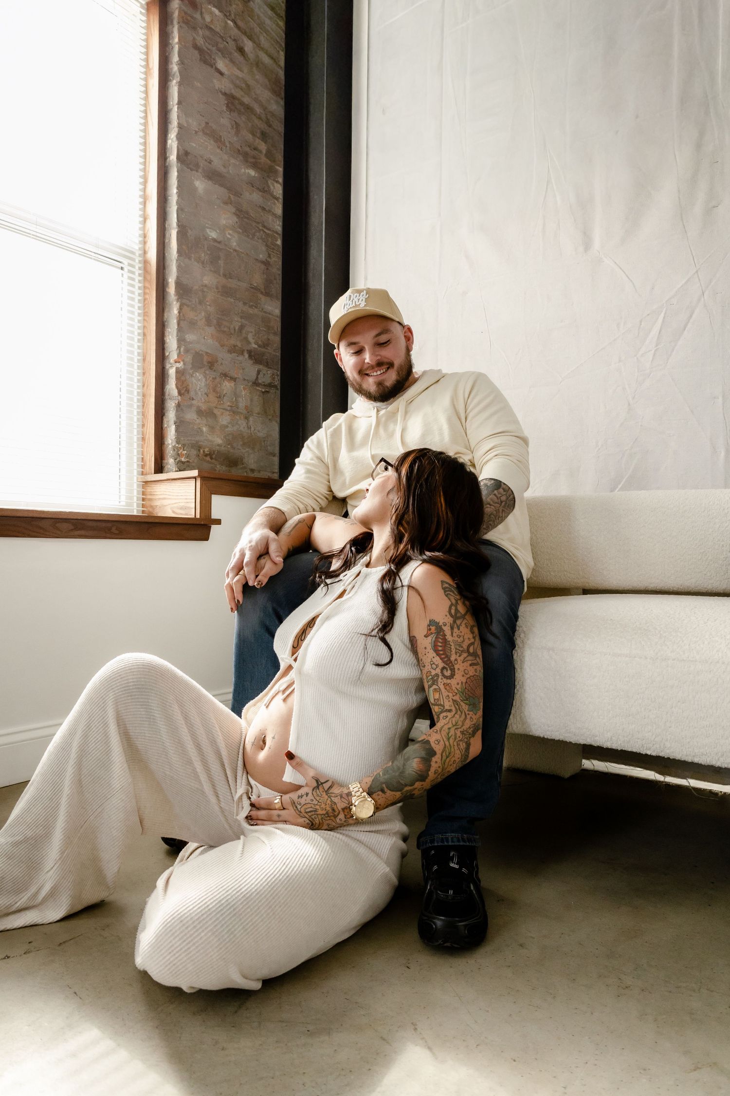 Intimate maternity photo session showing couple in white dress and black clothing sitting together on floor near window.