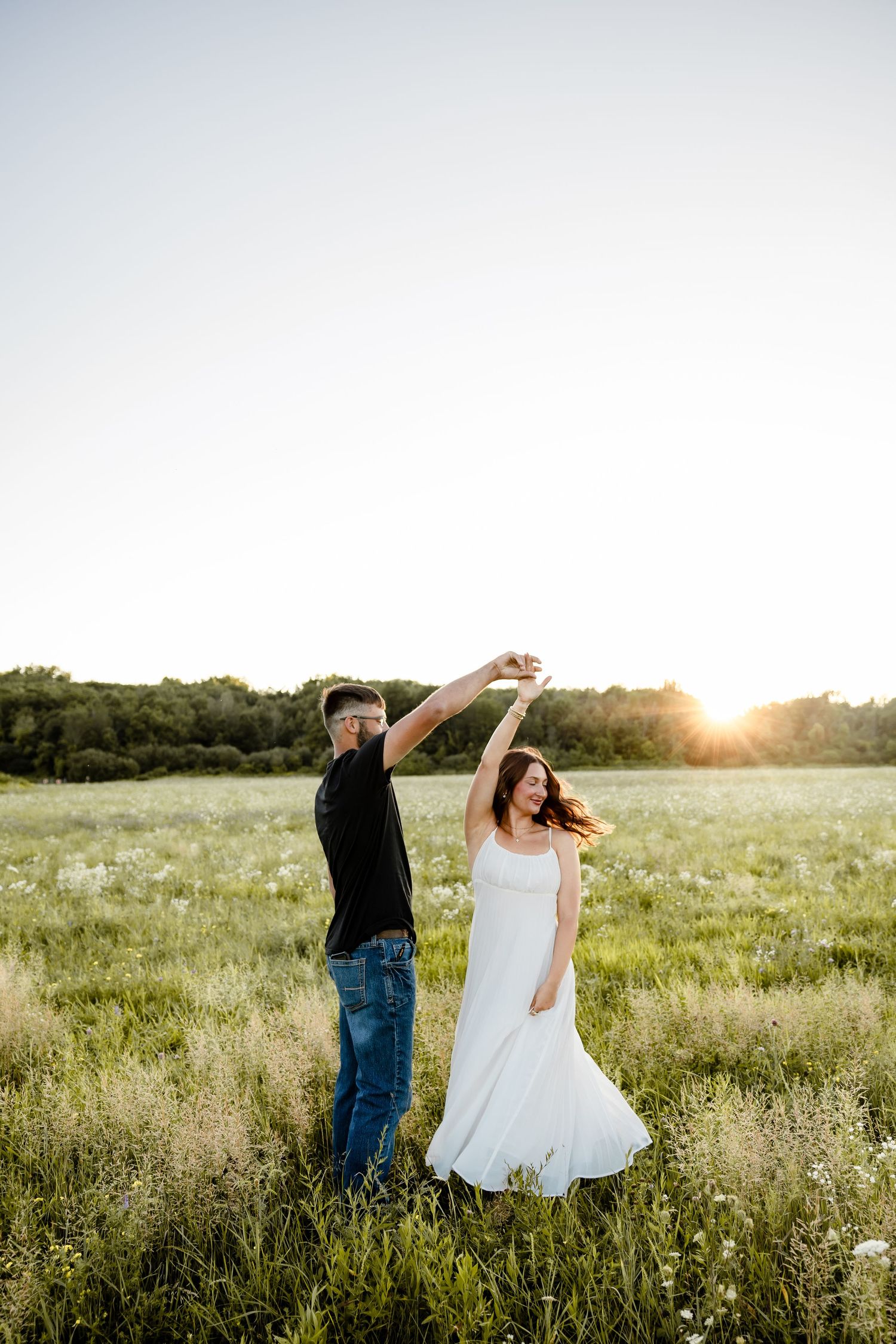 Couple dancing in a sunlit meadow, woman in white dress being twirled by man in black shirt and jeans at sunset.