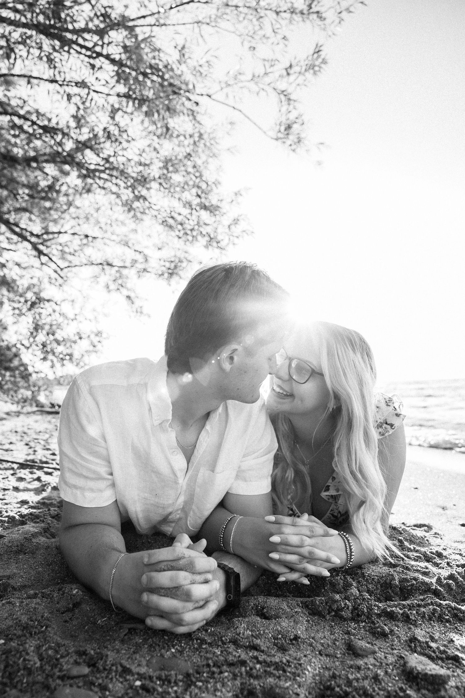 Black and white photo of a romantic couple lying on a sandy beach, sharing a tender moment under sun-dappled trees.