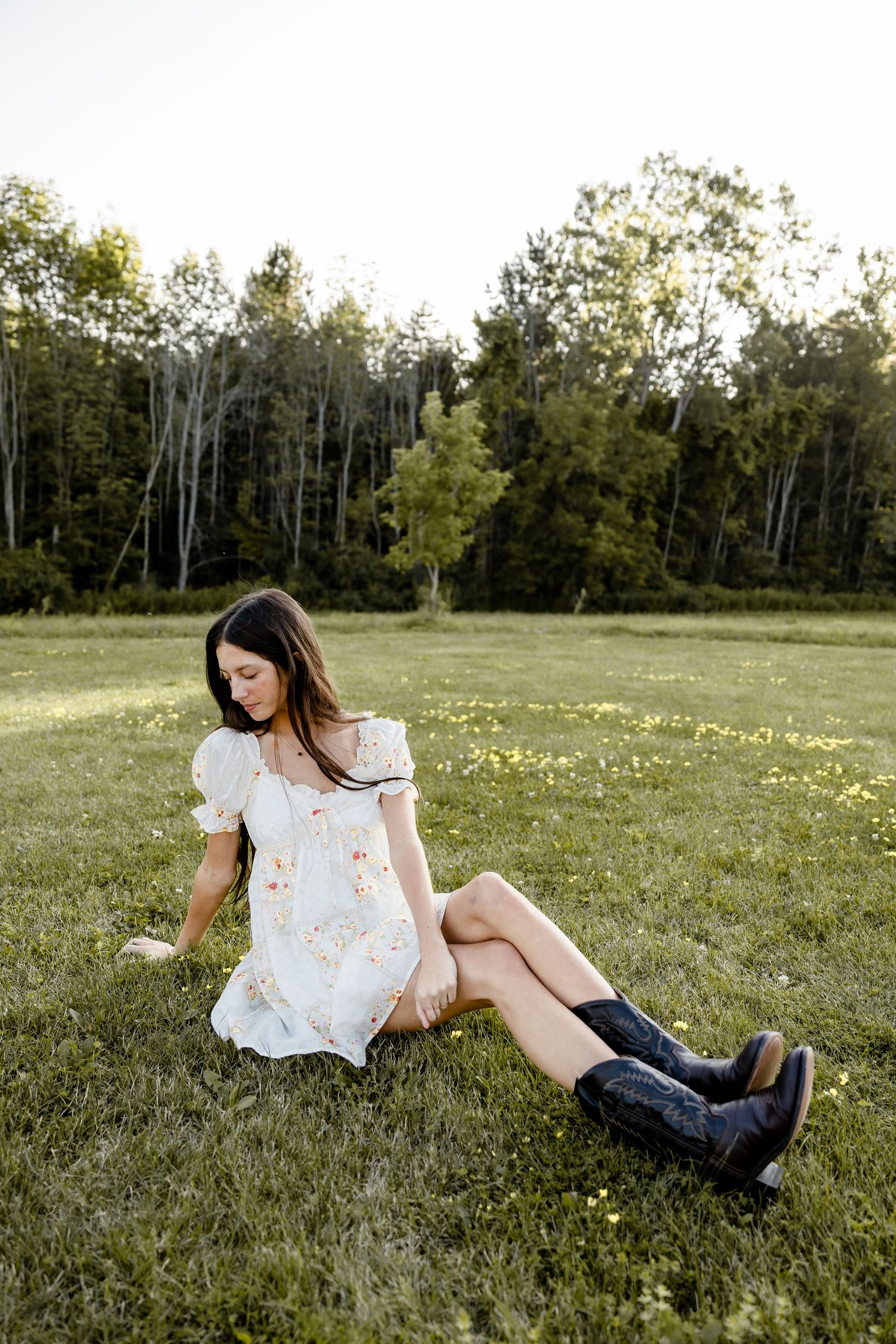 Young woman in white floral dress and cowboy boots sitting in a green meadow with trees in the background.