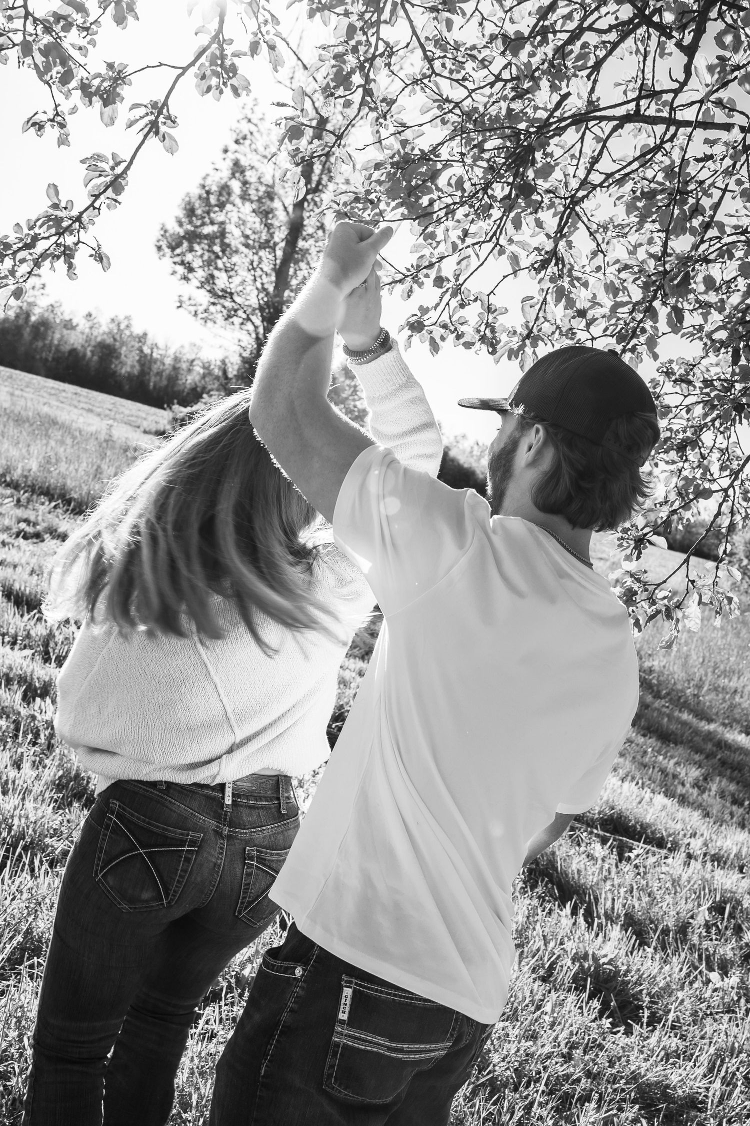 Black and white photo of a couple dancing outdoors near a tree in a sunny field, both wearing white tops and jeans.