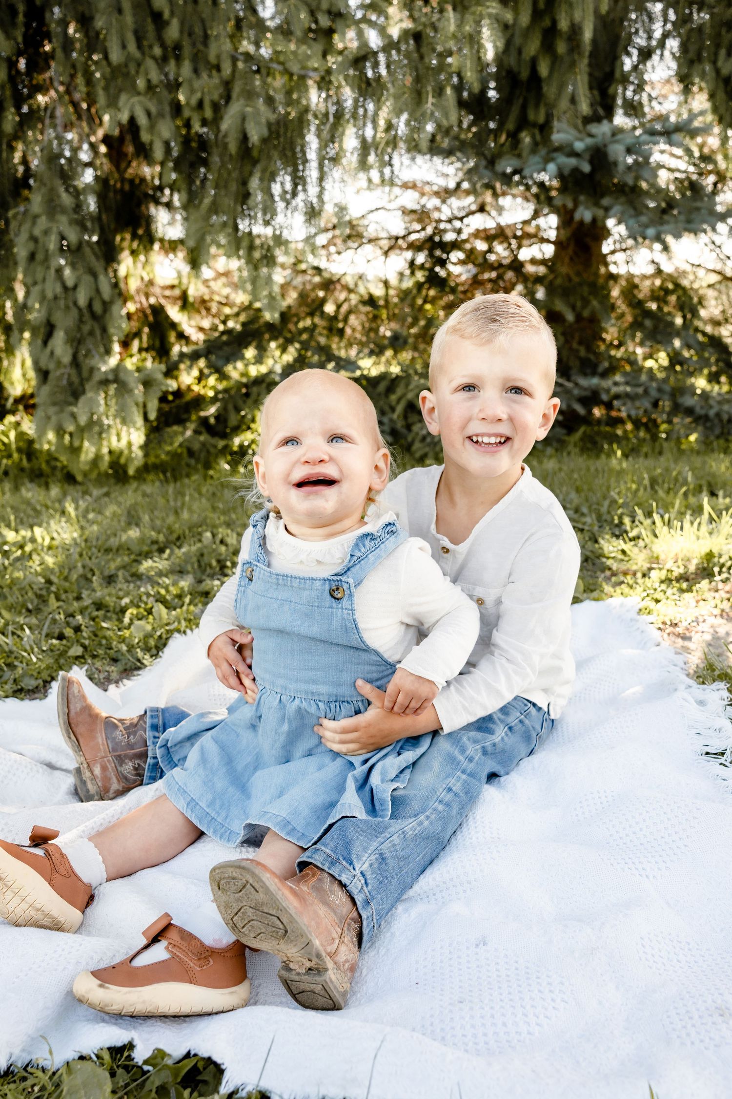 Two young siblings smile joyfully on a white blanket outdoors, baby in denim dress with older child in white shirt.