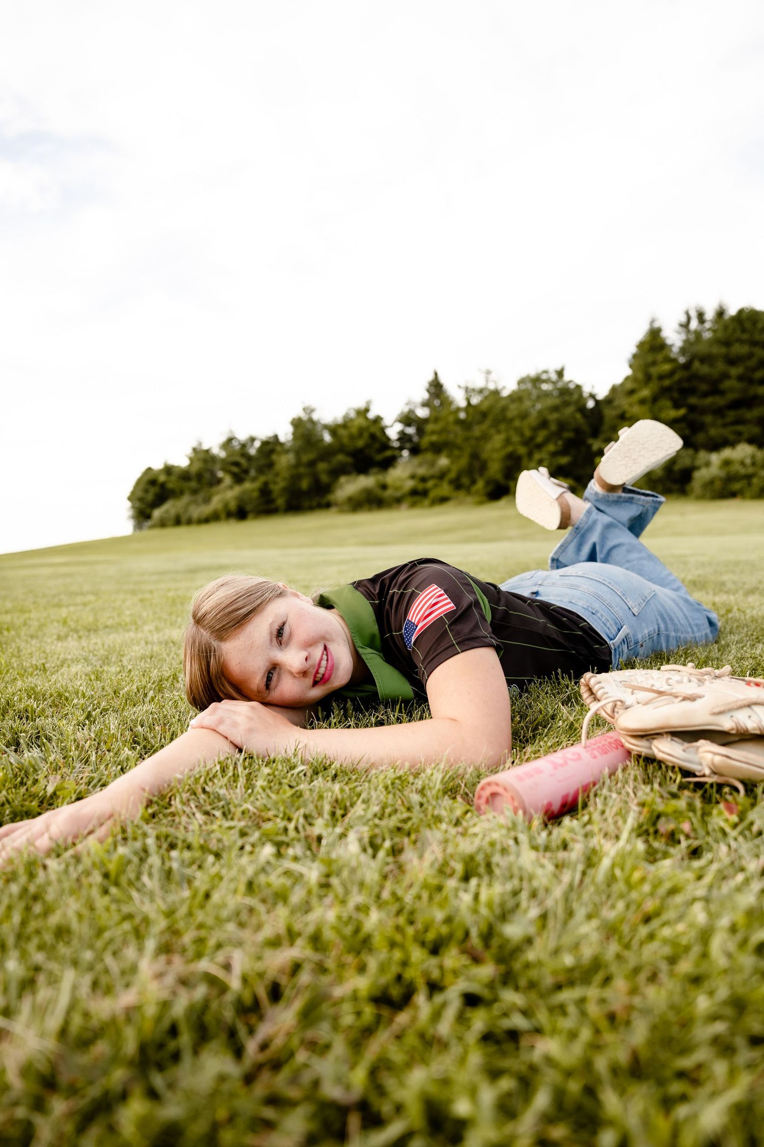 A smiling young girl lying on green grass in a park, wearing jeans and a black shirt, with a baseball glove nearby.