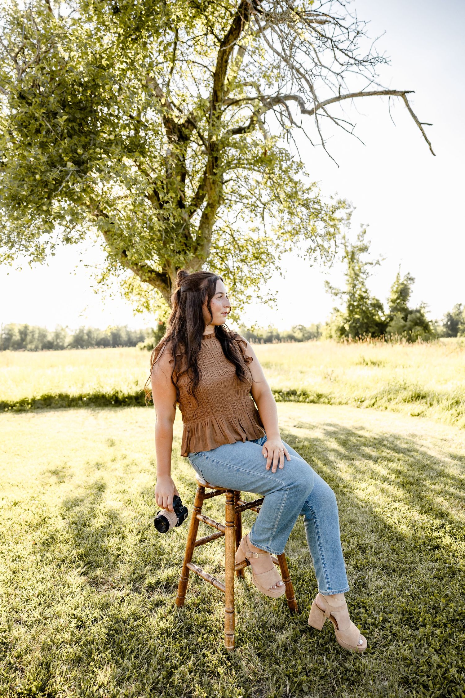 A young woman sits on a wooden stool under a tree in a sunny field, wearing jeans and a brown top.