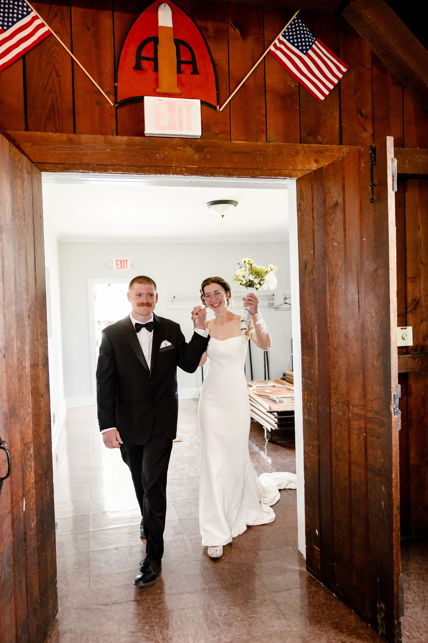 Bride and groom walk through wooden barn doors decorated with American flags at their wedding reception.
