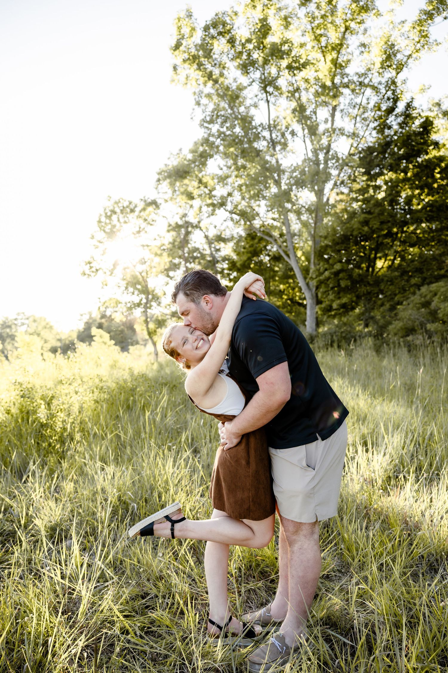 Couple sharing a romantic dip kiss in a sunlit green meadow surrounded by trees during golden hour.
