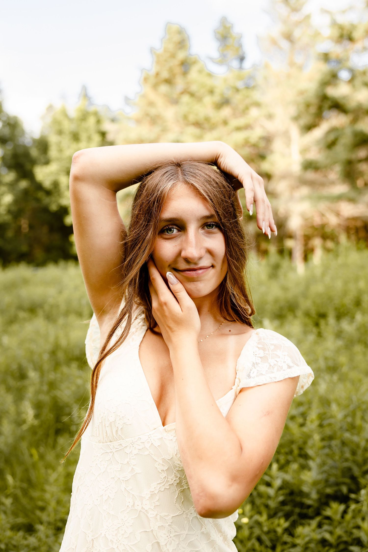 Young woman in white lace dress posing outdoors in green forest meadow, smiling with hand raised above her head.