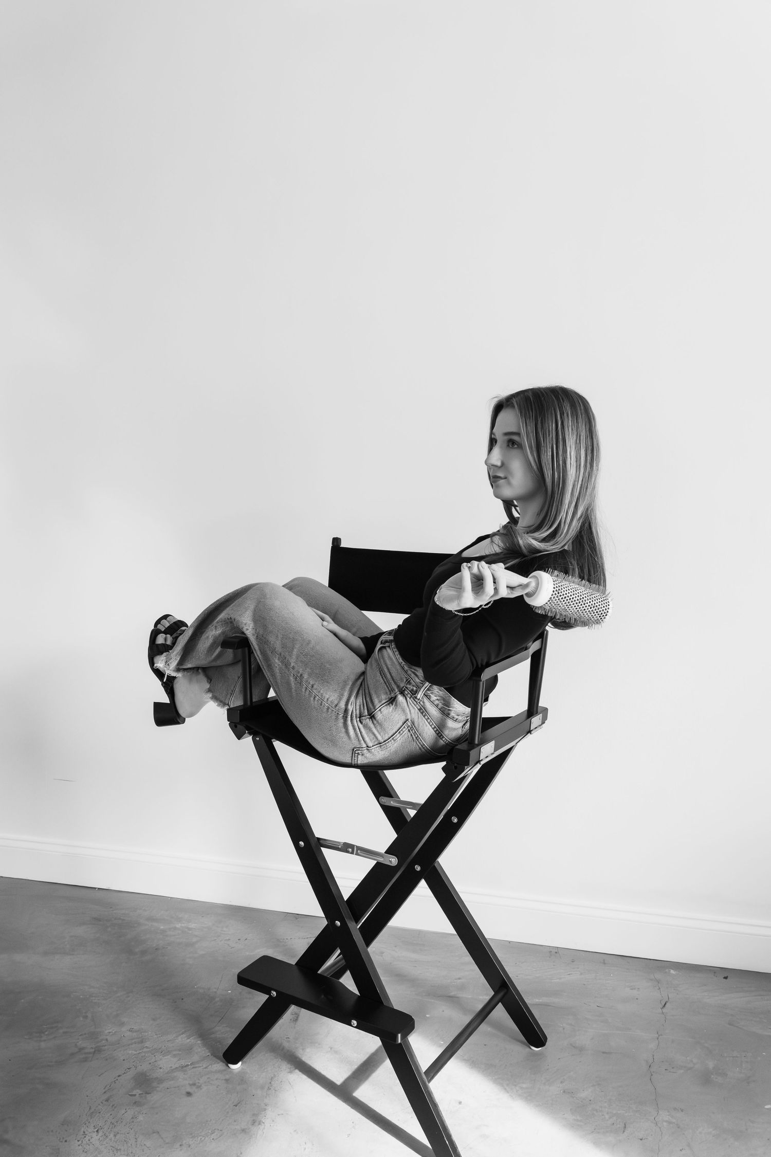 Black and white photo of a woman sitting in a director's chair with legs crossed, holding a drink.