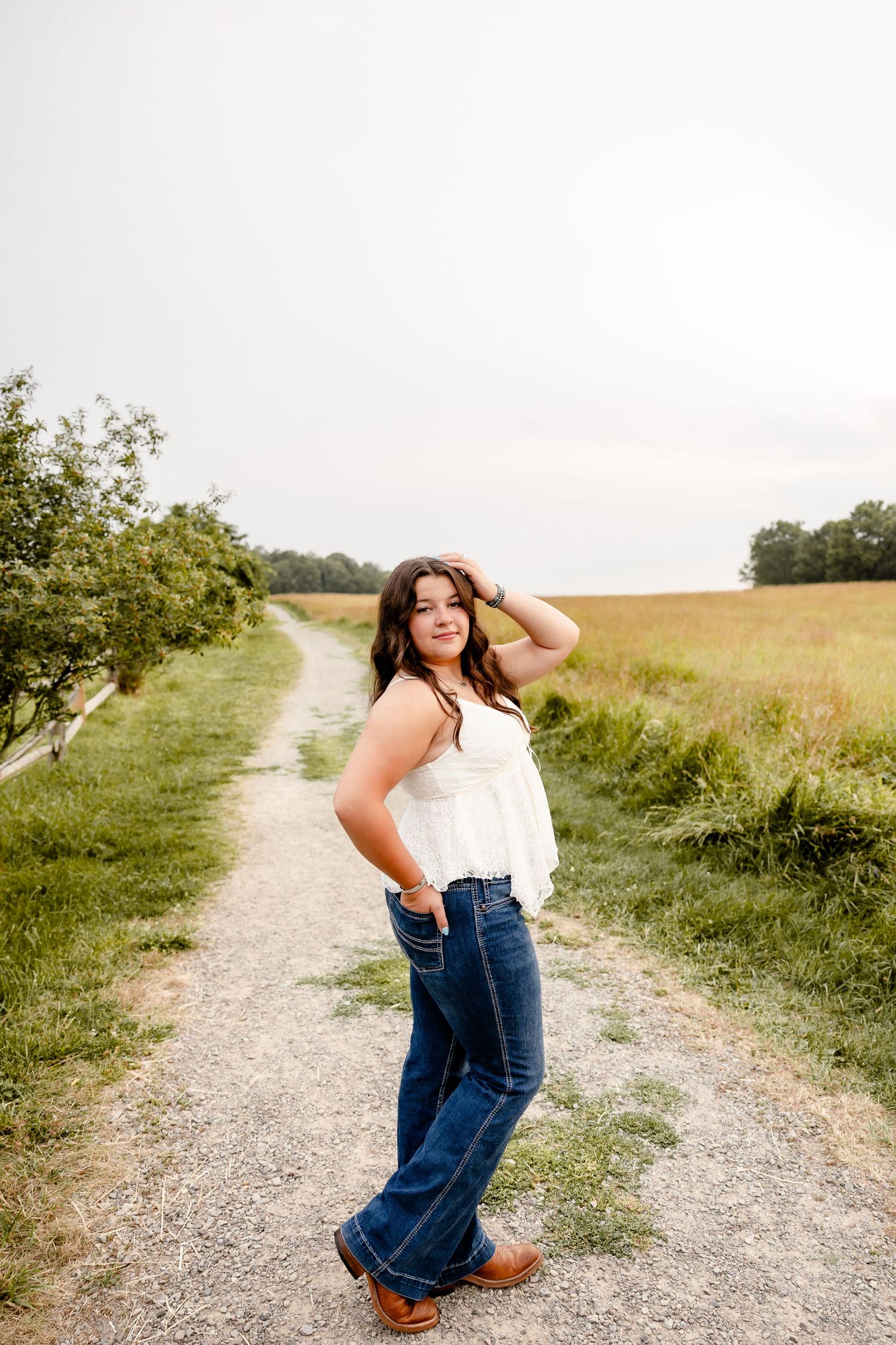 Young woman in white top, flare jeans, and cowboy boots poses on a rural dirt path surrounded by green fields.