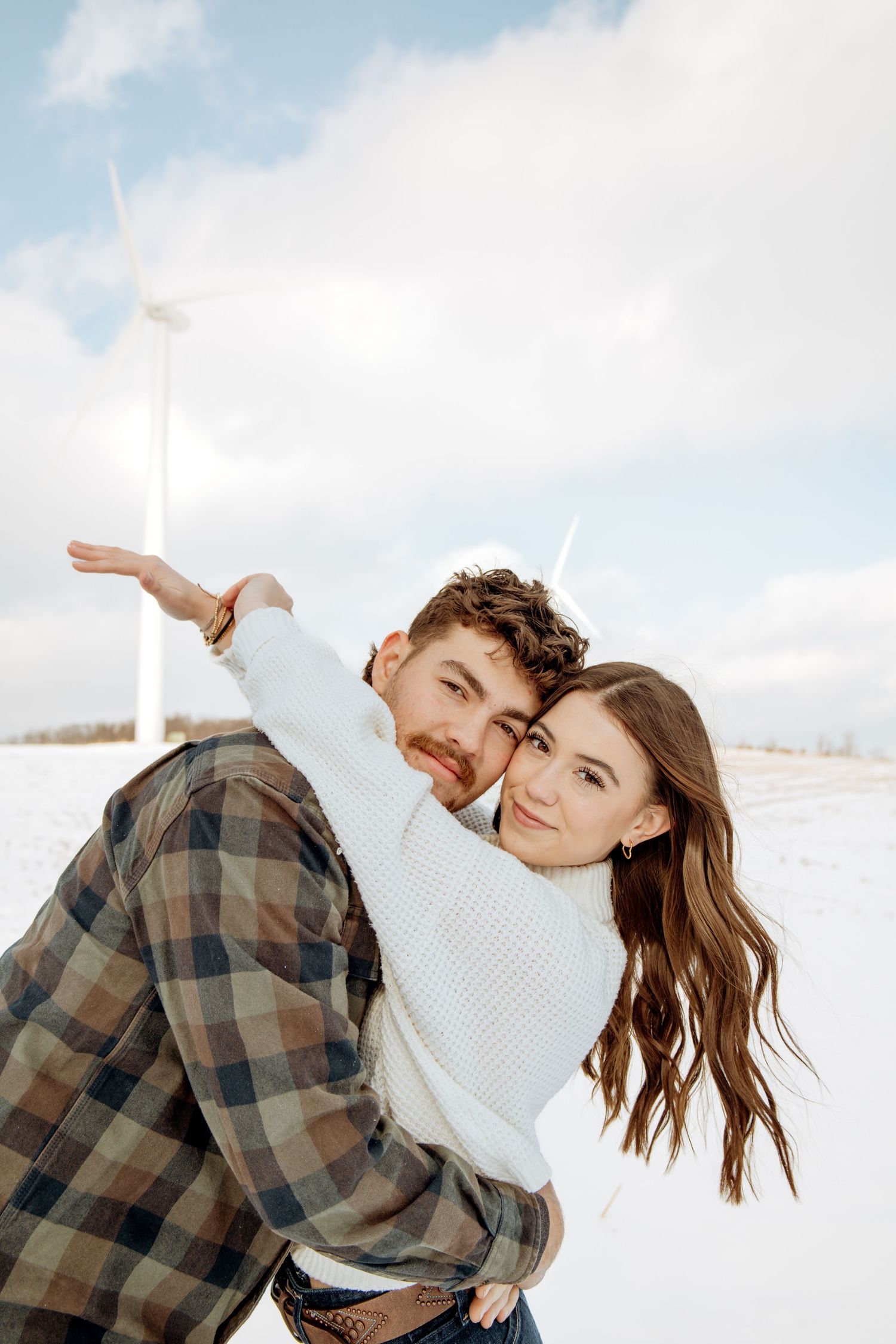 Couple embracing and smiling in a snowy field with wind turbines in the background on a cloudy day.