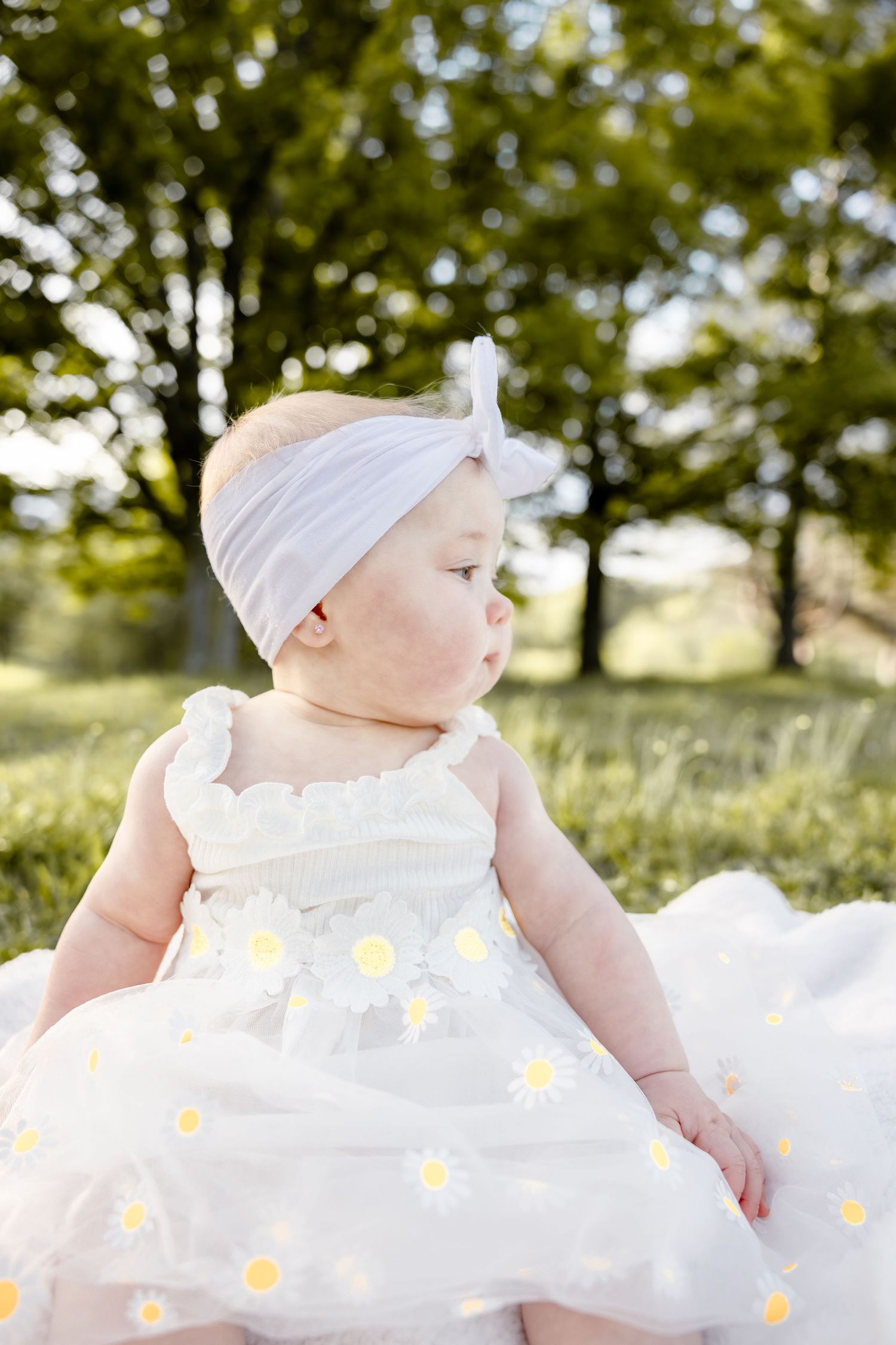 Adorable baby girl in white daisy dress and pink bow headband sitting outdoors in green park setting.