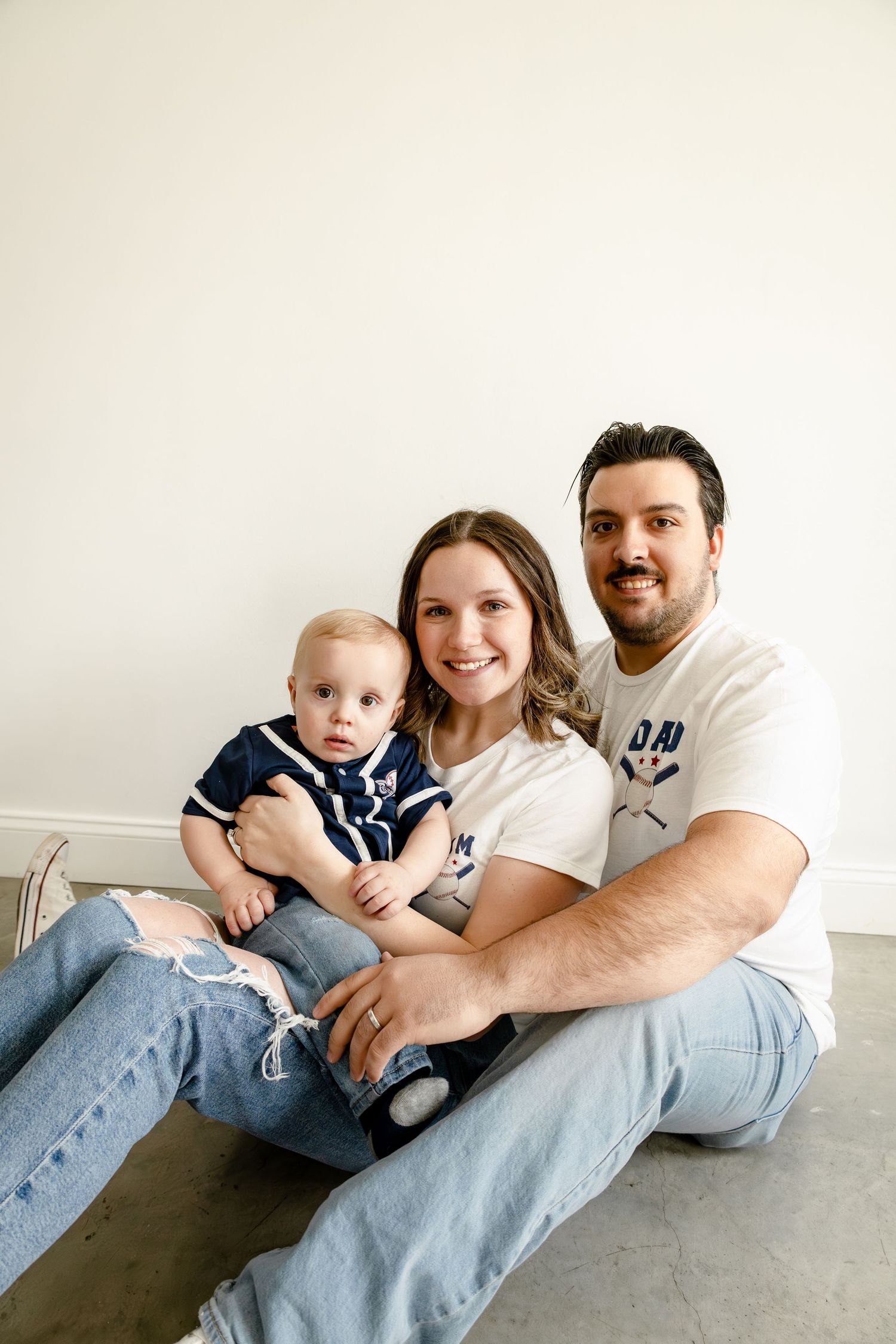 A young family with a baby boy sits on the floor smiling, wearing casual jeans and matching white and navy outfits.