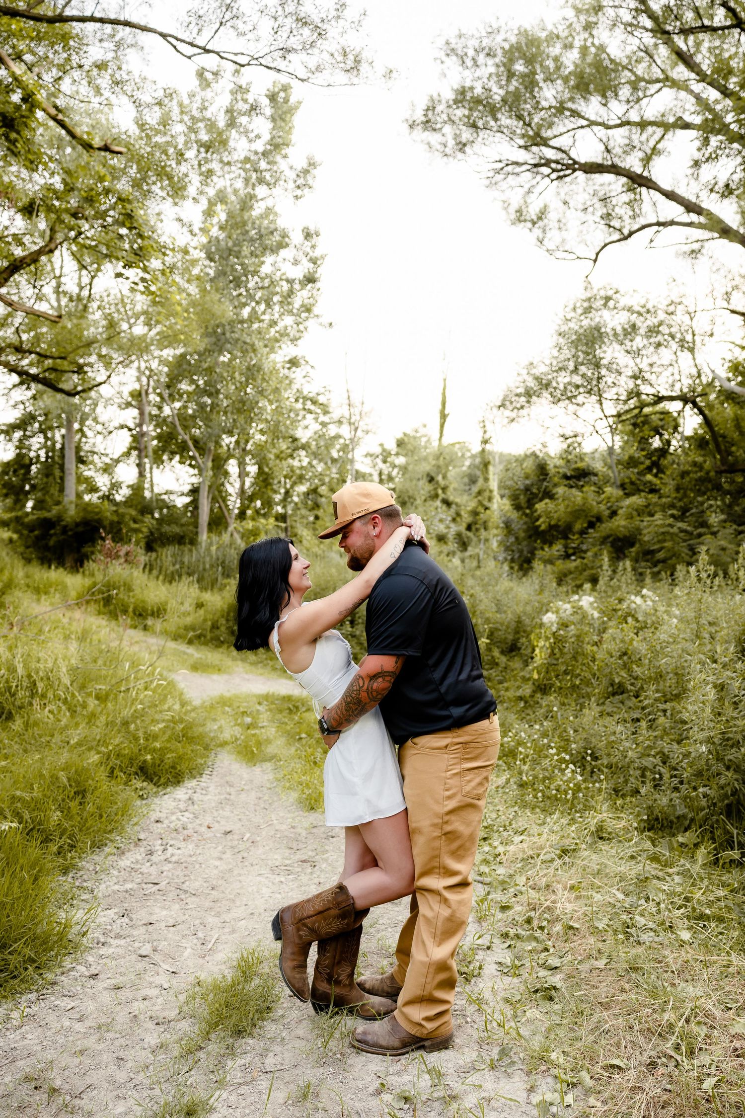 Couple embracing on a sandy trail surrounded by lush green trees, woman in white dress and boots, man in black shirt and hat.